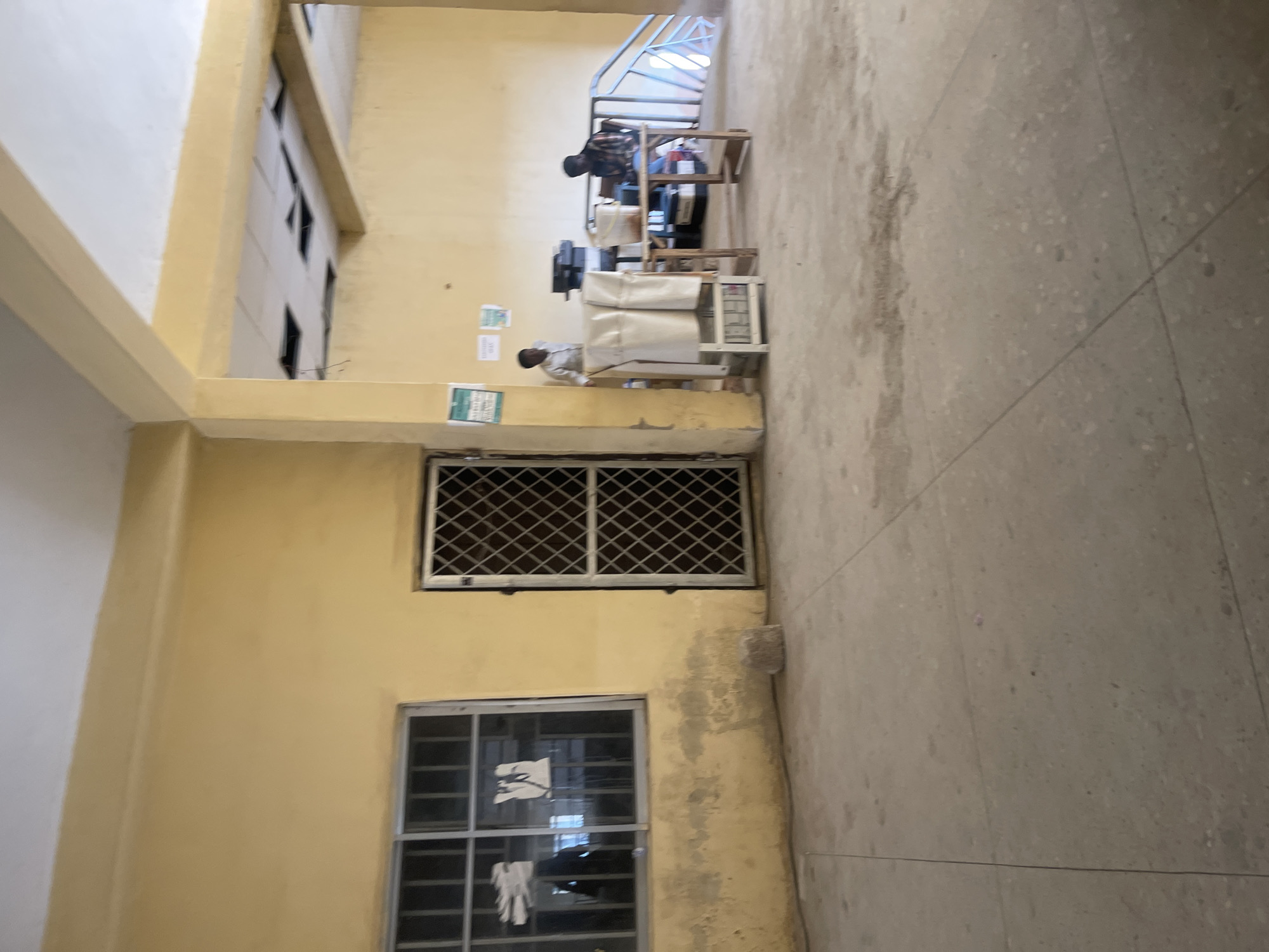 The image depicts an indoor scene, likely a public building, with a focus on a reception or waiting area. The walls are painted a pale yellow, and the floor is a tiled concrete. A barred window is visible on the left, with papers taped to the inside. To its right is a metal-barred door. In the background, two individuals are present. One, standing, appears to be a man in a light-colored shirt, possibly an attendant or official. He is near a wooden table laden with office equipment, including printers. Another individual, seated at the table, is wearing a plaid shirt and appears to be engaged with the equipment or documentation. A staircase with a light blue railing is visible further to the right. The ceiling shows exposed beams and some missing ceiling tiles, suggesting potential ongoing maintenance or a dated structure. The overall impression is of a functional, albeit somewhat austere, administrative space. The location of Rafin Par, Nigeria, places this scene within a specific geographical and cultural context, though no specific visual cues within the image directly confirm this location beyond the provided information.