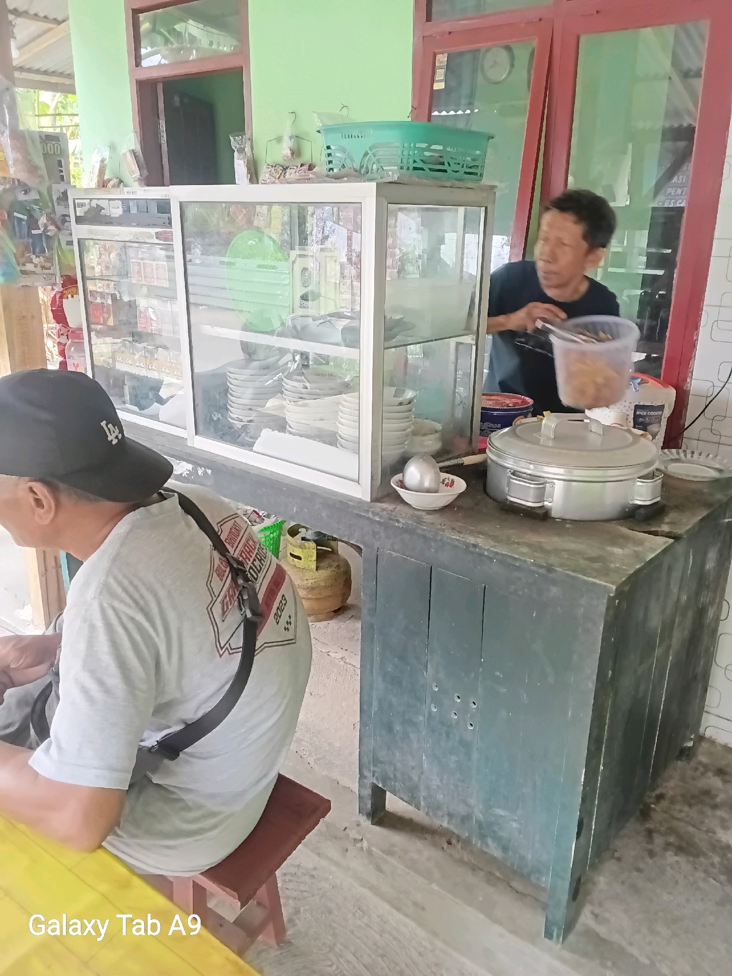 In a street-side stall in Karangsoko, Indonesia, a man in a dark t-shirt is preparing food. He is standing behind a glass display case filled with various items, including what appear to be snacks and beverages. In front of the display case, on a dark grey cabinet, sits a large, silver, lidded pot, likely for keeping food warm. Next to it, a small white bowl contains a metallic-colored object, possibly an egg. A clear plastic container, held by the man, is filled with small, brown food items, possibly fried snacks. A second person, wearing a grey t-shirt and a black baseball cap, is seated on a wooden stool, facing away from the camera, looking towards the stall. Behind them, a yellow gas cylinder is visible. The stall is simple, with a green painted wall and a red-framed window in the background. The overall scene depicts a typical food stall environment.