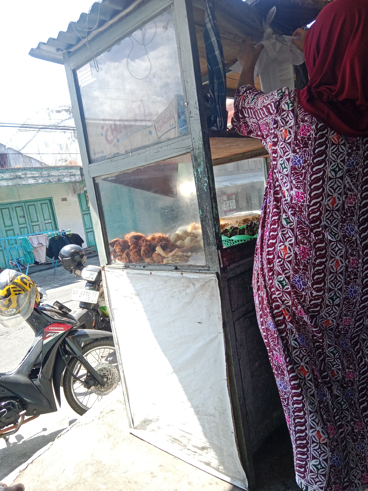 In Ringinanom, Indonesia, a street food vendor stands behind a glass-fronted cart. The vendor, wearing a maroon hijab and a patterned batik dress, is placing an item into a white plastic bag. The cart displays fried food items, possibly chicken, behind the glass. A motorcycle is parked to the left of the cart, with two helmets placed on its handlebars. In the background, a clothesline with laundry is visible, along with buildings and a fence. The scene is illuminated by bright sunlight, casting shadows on the ground.