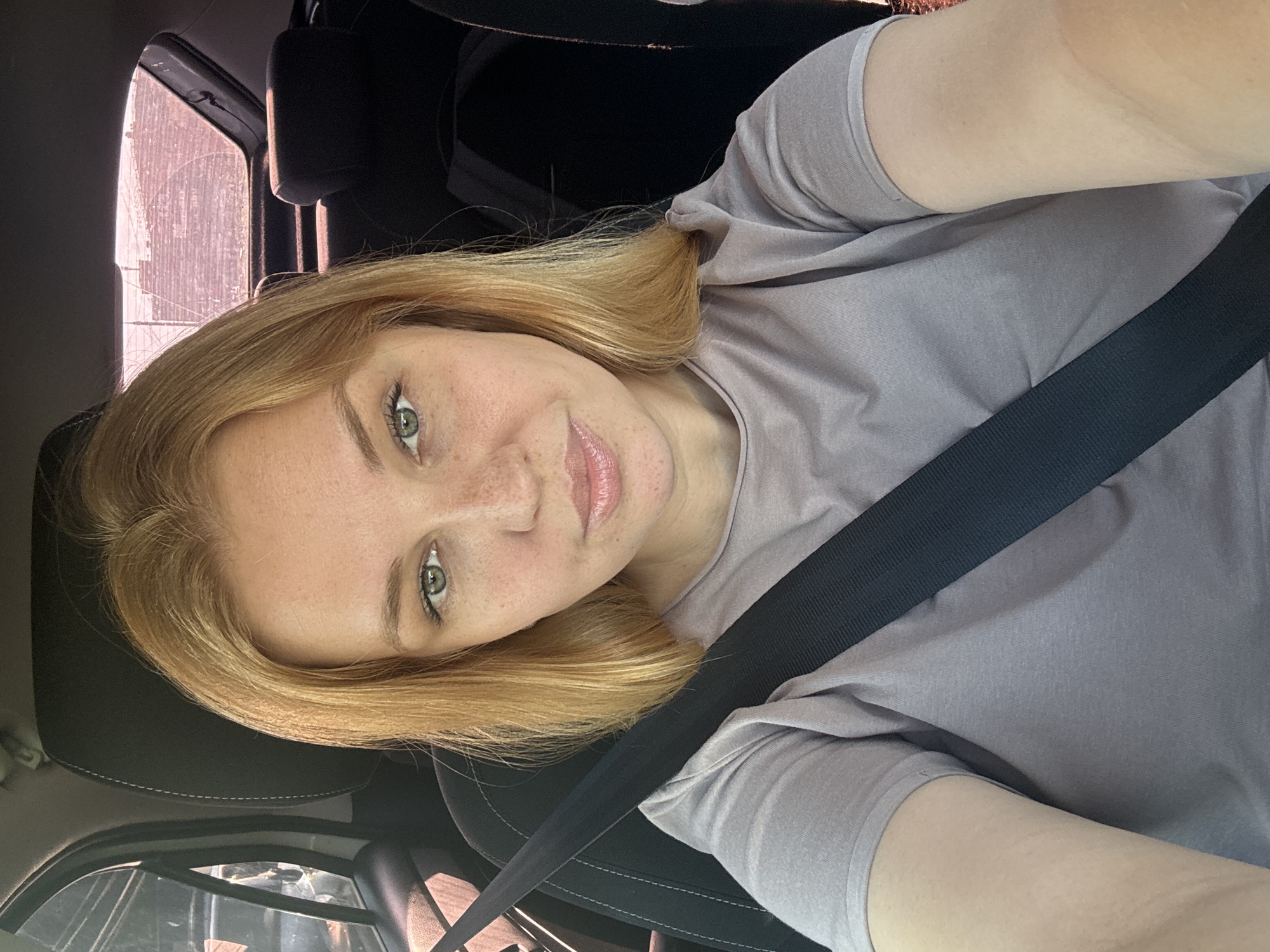 A young woman with blonde hair and green eyes is sitting in the driver's seat of a car. She is wearing a light gray t-shirt and her seatbelt is fastened. The car appears to be parked or stopped, as indicated by the interior view and the ambient lighting which suggests it is daytime. The background shows the interior of the car, including headrests and windows, with a glimpse of what appears to be an urban or suburban setting outside the windows. The image is a selfie taken by the woman. There is no information in the image to suggest a specific location within the city of Zhukovo, Russia.