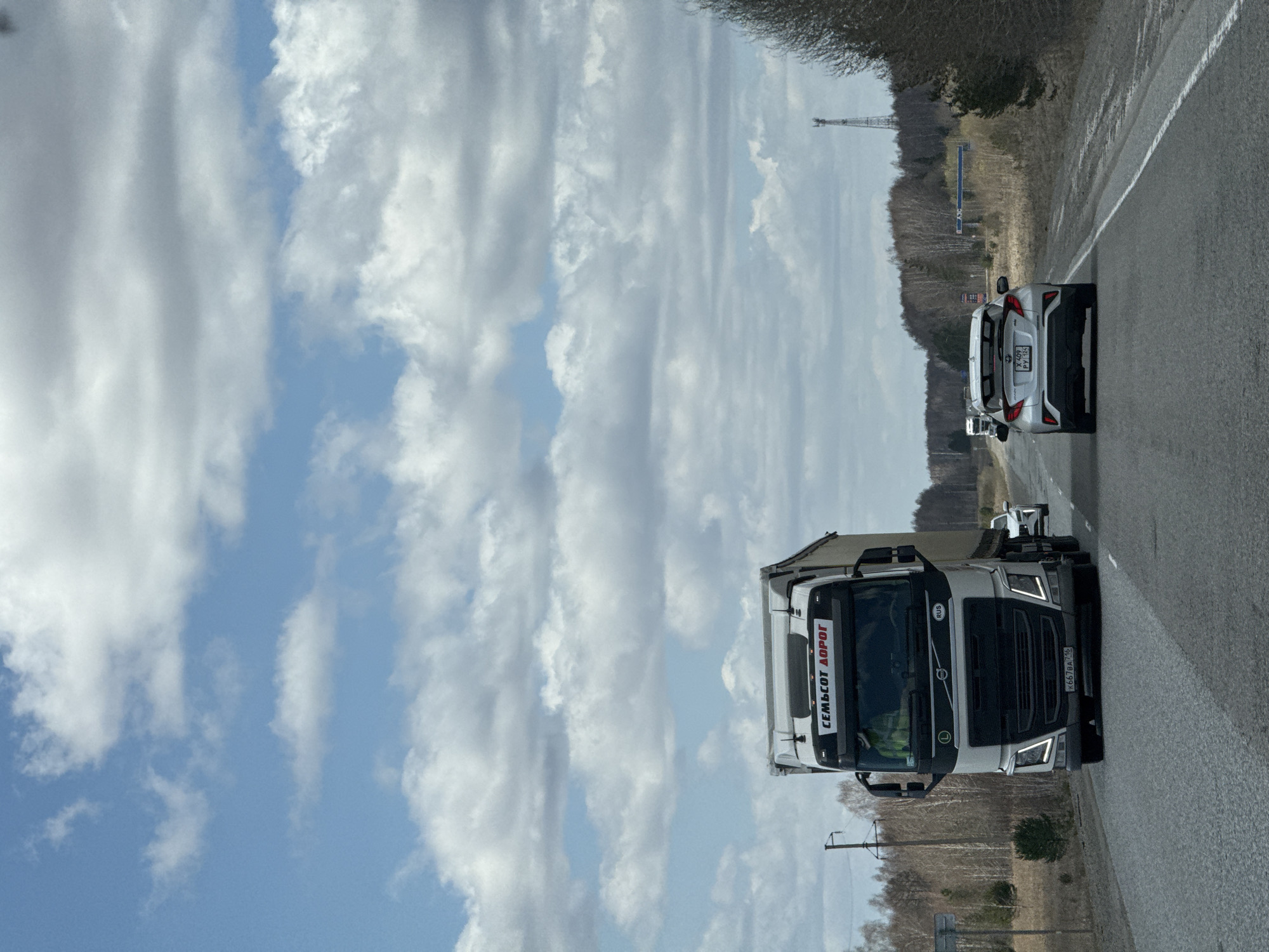 The image shows a road with a white truck and a white car driving on it. The truck has a Russian license plate "K667BA 146". The word "СЕМЬСОТ ДОРОГ" is visible on the front of the truck, which means "Seven Hundred Roads" in Russian. There are trees on both sides of the road. The sky is cloudy with patches of blue. There is a tall tower in the distance. Given the truck's writing "СЕМЬСОТ ДОРОГ" and the license plate, the location is likely to be Russia. However, there is insufficient data to pinpoint the exact location as Mar'evka, Russia.