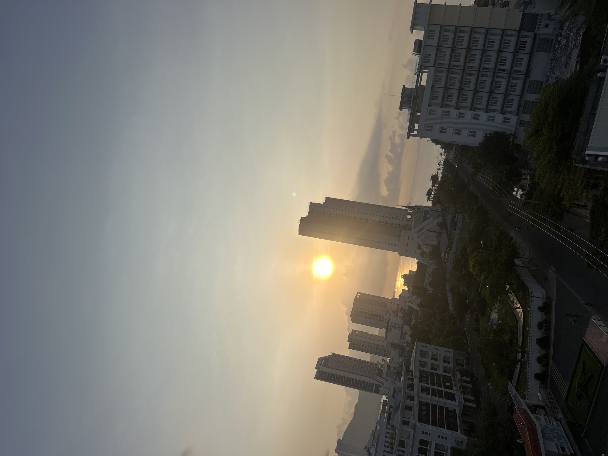 The image depicts an urban scene at sunset in Bac Nha Trang Ward, Vietnam. Several high-rise buildings dominate the view, silhouetted against the setting sun. A multi-story structure with numerous windows stands in the top right corner, suggesting a residential or commercial building. Other buildings of varying heights populate the landscape, some appearing newer and others older. In the distance, a glimpse of the ocean is visible, reflecting the sunlight. The sky displays a gradient from light gray to orange, indicating the transition from day to night. A road winds through the scene, flanked by trees and greenery.