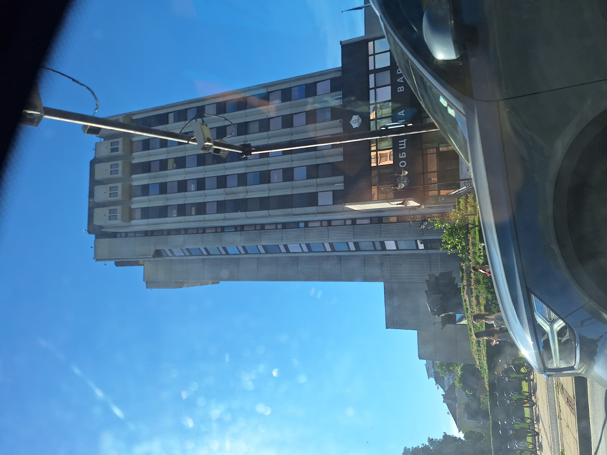 The image captures a view from a vehicle, looking towards a tall, Brutalist-style concrete building under a clear blue sky. The building is the Varna Municipality building, located in Varna, Bulgaria. In the foreground, the hood and headlight of a dark grey car are visible. To the left of the building, a row of green bicycles is parked, suggesting a bike-sharing station. Further to the left and behind the bicycles, some trees and residential buildings with red roofs can be seen. In front of the main building, there is a sculpture and a tiered garden with flowering plants. Three women are walking in the garden area. The lower section of the Varna Municipality building features large windows and signage that reads "ОБЩИНА ВАРНА" (Municipality of Varna). A street lamp and a surveillance camera are mounted on a pole in front of the building. The overall scene depicts an urban environment on a sunny day.