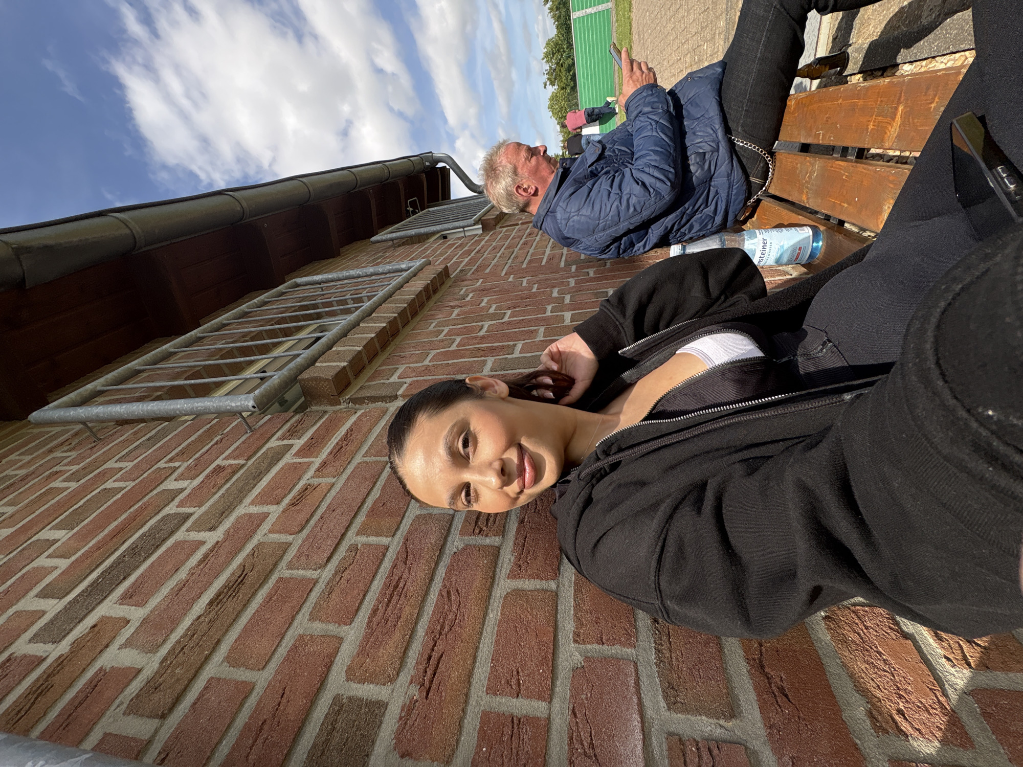 This outdoor image, captured in Wegberg, Germany, depicts a young woman and an older man seated on a wooden bench in front of a brick building under a clear blue sky with scattered white clouds.
In the left foreground, a young woman with dark hair pulled back offers a subtle smile directly at the viewer. She is dressed in a black zip-up hoodie over a white top and dark leggings, with her right hand resting near her neck. A black smartphone lies on her lap.
To her right, an older man with fair hair is seated in profile, looking down at and operating his smartphone. He is dressed in a dark blue quilted jacket, dark trousers, and has a metal chain attached to his belt loop. A clear plastic bottle of Warsteiner water stands on the wooden bench between the two individuals, with another dark bottle partially visible beneath the man's seat.
The brick building behind them features a wall of varying red and brown bricks with light mortar lines, topped by a metal railing and a grey rain gutter system beneath a dark wooden soffit. Beyond a paved area in the midground, the distant background reveals a green fence, likely bordering a sports field, with trees and small figures, including one in a red shirt, visible under the bright blue sky.