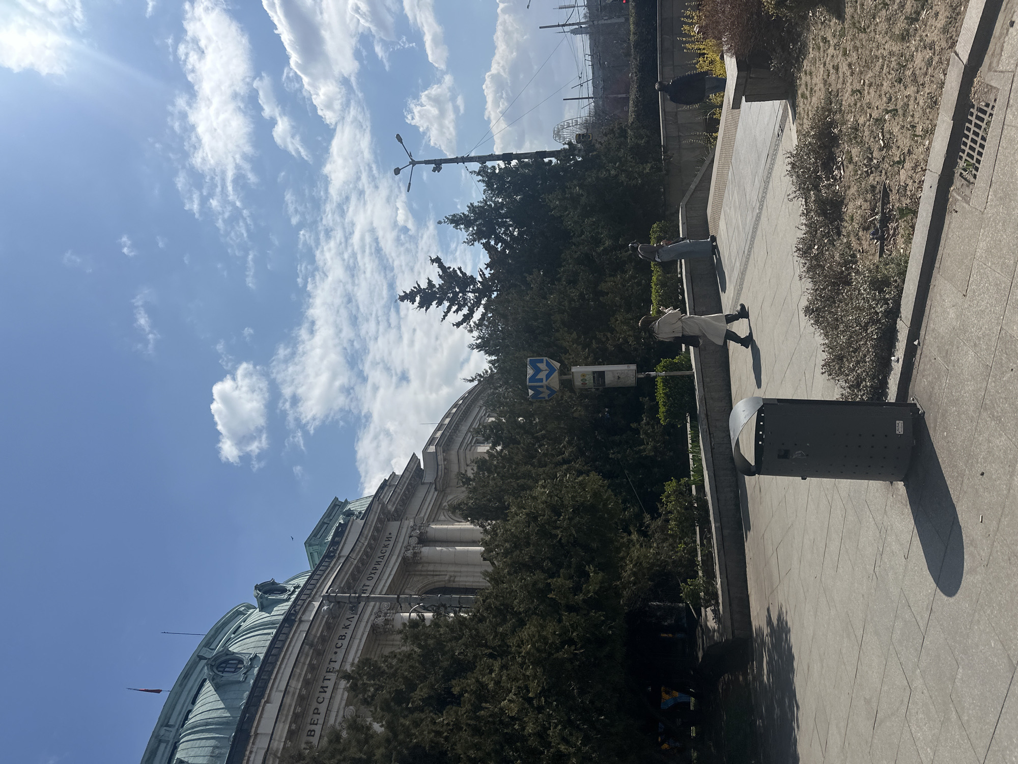 The image depicts an outdoor urban scene on a bright, sunny day in Sofia, Bulgaria, specifically showcasing the Sofia University "St. Kliment Ohridski."
The prominent structure on the left is the Sofia University "St. Kliment Ohridski," identified by the inscription "УНИВЕРСИТЕТ • СВ. КЛ. ОХРИДСКИ" on its classical facade. The building features a large, verdigris-colored dome with multiple oculus windows and a red flag staff. The facade below the dome is adorned with columns and ornate carvings. Numerous dark green evergreen trees partially obscure the lower and mid-sections of the university building.
The foreground consists of a paved pedestrian area made of light-colored tiles. A dark grey, cylindrical public waste bin with a curved top is positioned in the lower left quadrant, casting a distinct shadow to its right. A dry, sparse patch of earth and low-lying vegetation is visible to the right of the bin and in the mid-ground.
Three individuals are present. One person, wearing a light-colored long coat and dark trousers, is walking from left to right on the paved surface in the foreground. A second individual, dressed in a light-colored top and jeans, stands further back on a raised concrete platform, facing away from the camera. A third individual, wearing a dark jacket, stands to the right, partially obscured by vegetation and a concrete barrier. No direct interactions are discernible between the individuals.
A blue square sign with a white stylized "MM" symbol, characteristic of the Sofia Metro, is positioned in the mid-ground, indicating a nearby metro station. In the mid-ground to the right, a tall, multi-headed streetlamp is visible. Further in the distant background, a large Ferris wheel and overhead power lines are present under a sky with scattered white cumulus clouds and strong sunlight.