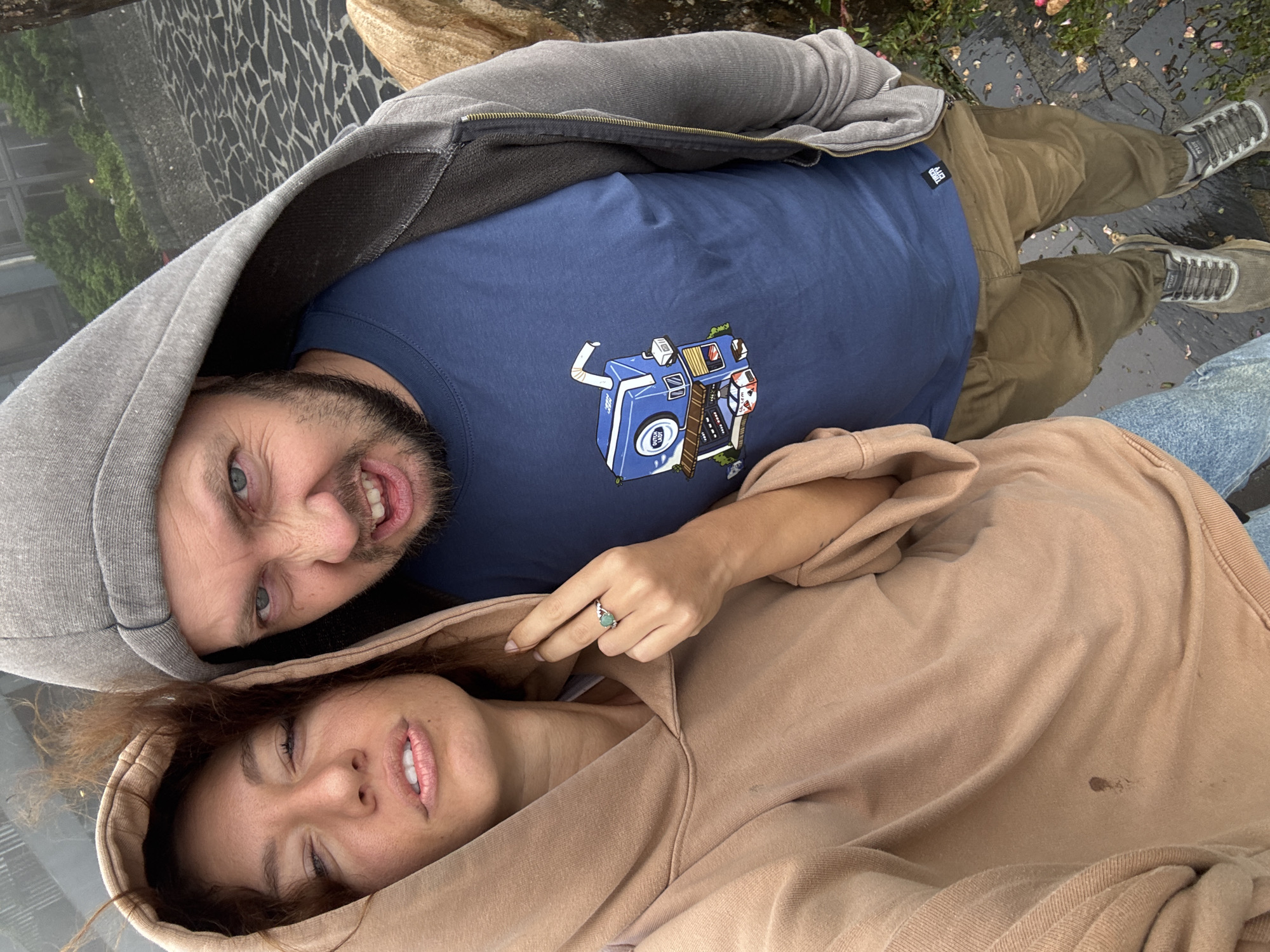 A man and a woman are lying down outside. The man, on the upper left, is wearing a gray hooded sweatshirt, a navy blue t-shirt with a graphic design, and khaki pants. He is smiling widely, showing his teeth. The woman, on the lower right, is wearing a tan hooded sweatshirt and has a ring on her finger. They are lying on what appears to be a glass surface that reflects the surrounding environment. The ground shows a pattern of irregular black shapes on a gray background, and there are trees and building reflections visible on the surface. They are in Binh Lu, Vietnam.