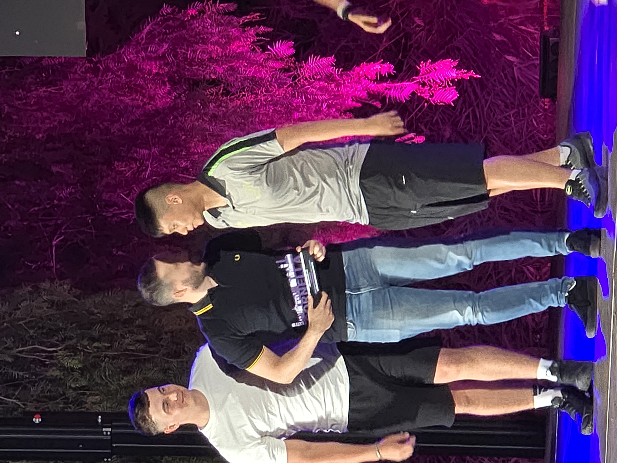 This image depicts an awards ceremony held outdoors, likely in Cornellà de Llobregat, Spain, given the event's branding. Three men are the primary subjects on stage. The man on the left, wearing a white t-shirt and black shorts, is looking to his left. In the center, a man in a black polo shirt with yellow trim and blue jeans is holding an award. He is looking at the man to his right. The man on the right, dressed in a grey polo shirt with green accents and black shorts, is also looking towards the man in the center. The background features lush greenery illuminated with vibrant pink lighting, creating a dramatic ambiance. The stage floor is reflective and illuminated with blue light.
