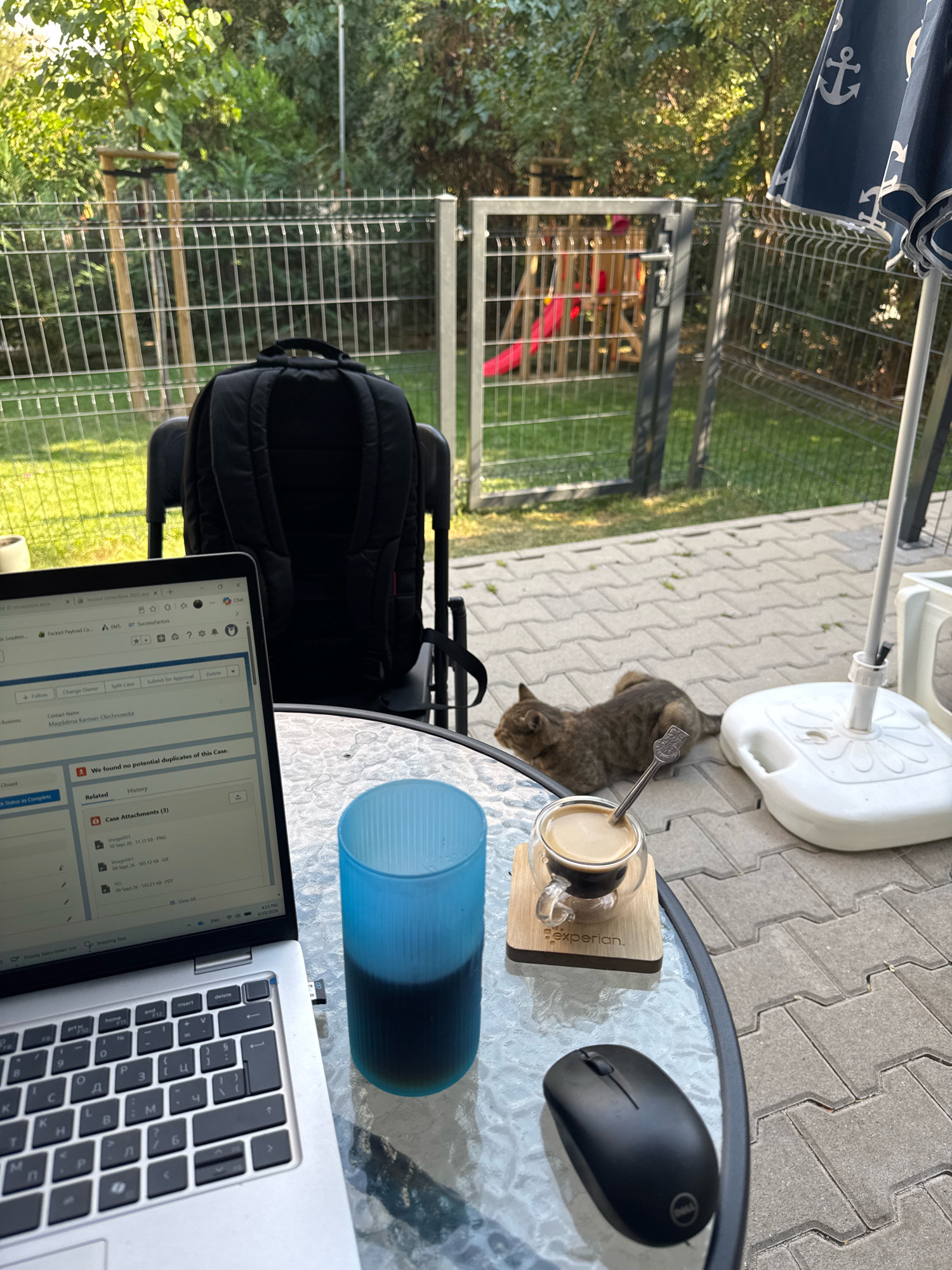 A dappled sunlight filters through lush green foliage onto a patio paved with light gray interlocking bricks. In the foreground, a silver laptop rests on a round glass table, its screen displaying a document with text. To the right of the laptop, a black computer mouse sits on the table. Adjacent to the mouse, a clear glass cup filled with dark coffee rests on a wooden coaster with "experian." printed on it. To the left of the laptop, a tall, fluted blue glass contains a dark liquid. In the background, a brown tabby cat is lying on the patio. Behind the cat, a black backpack is placed on a dark chair. A wire fence encloses a grassy backyard area with a wooden play structure visible through an open gate. A large navy blue umbrella with white anchor patterns is partially visible on the right side of the frame, attached to a white base. The environment suggests an outdoor setting, possibly a backyard or a patio, bathed in natural light. The presence of a laptop, mouse, and coffee implies a person working or relaxing outdoors. The general scene appears to be set in a residential area in Sofia, Bulgaria, given the characteristic fencing and paving stones often found in European urban backyards.