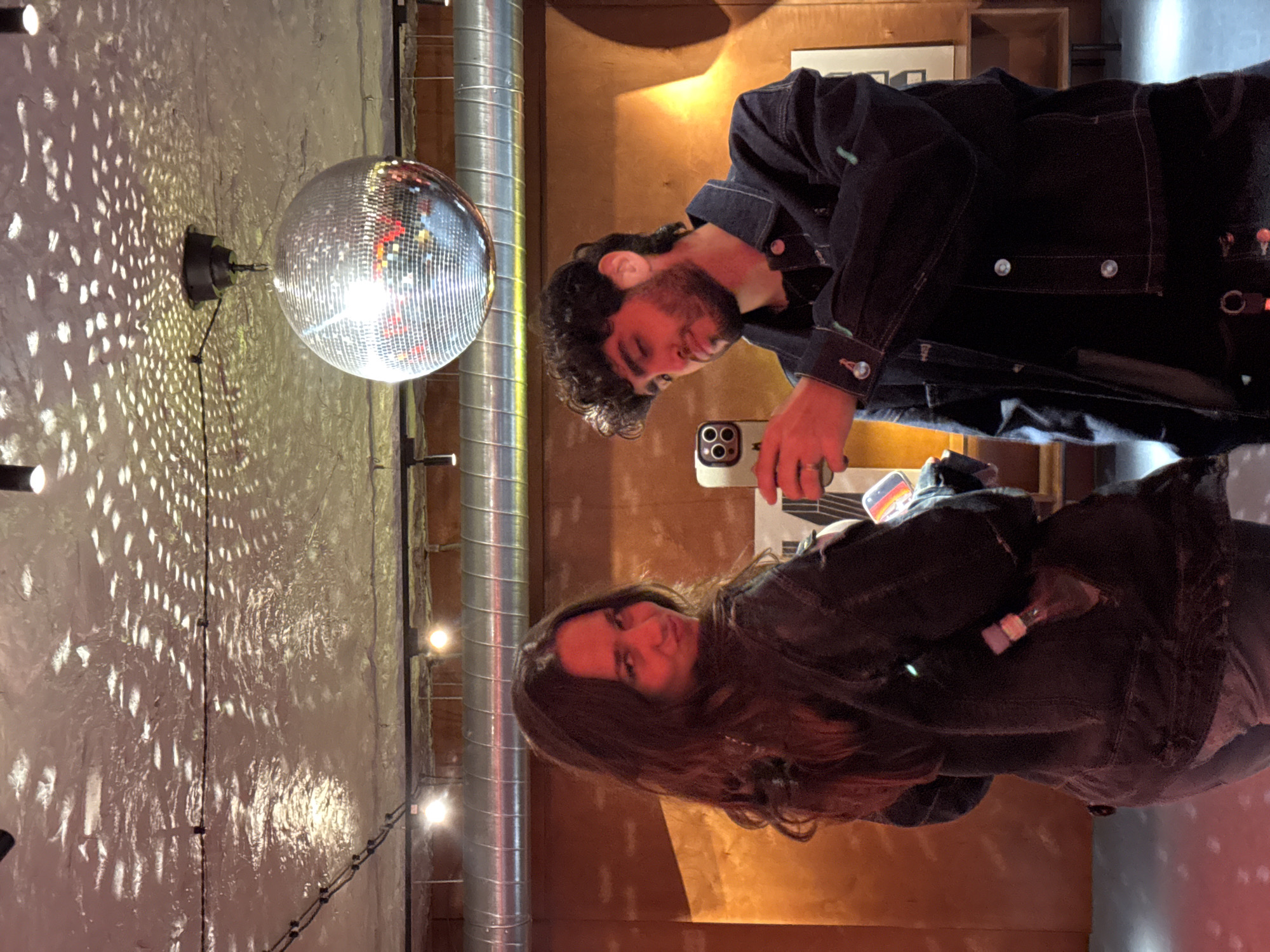 This image, captured indoors in Yerevan, Armenia, depicts a casual social scene featuring a man and a woman in ambient lighting. The scene is observed as if the original image has been rotated 90 degrees counter-clockwise to achieve a portrait orientation.
On the right side of the frame, a man with dark, curly hair and a beard is visible, wearing a dark denim jacket. He is holding a smartphone, likely an iPhone with multiple cameras, in front of him, suggesting he might be taking a photo or video. Rings are visible on his left hand. His gaze is directed towards the phone screen.
On the left, a woman with long, dark hair also wears a dark denim jacket. She looks towards the camera with a neutral to slightly smiling expression. An object, possibly a small bottle or flask, is tucked into her jacket pocket.
The background features a textured wall, possibly concrete or plaster, bathed in warm light. A prominent disco ball hangs from the upper left, casting numerous small, bright, dappled light patterns across the wall. Metallic pipes or ducts run vertically along the wall, contributing to an industrial-chic aesthetic. String lights with individual warm-toned bulbs are visible in the background, enhancing the cozy, evening atmosphere. The overall setting appears to be a bar, club, or a similar entertainment venue. No discernible text is visible in the image. The lighting suggests it is evening or nighttime.