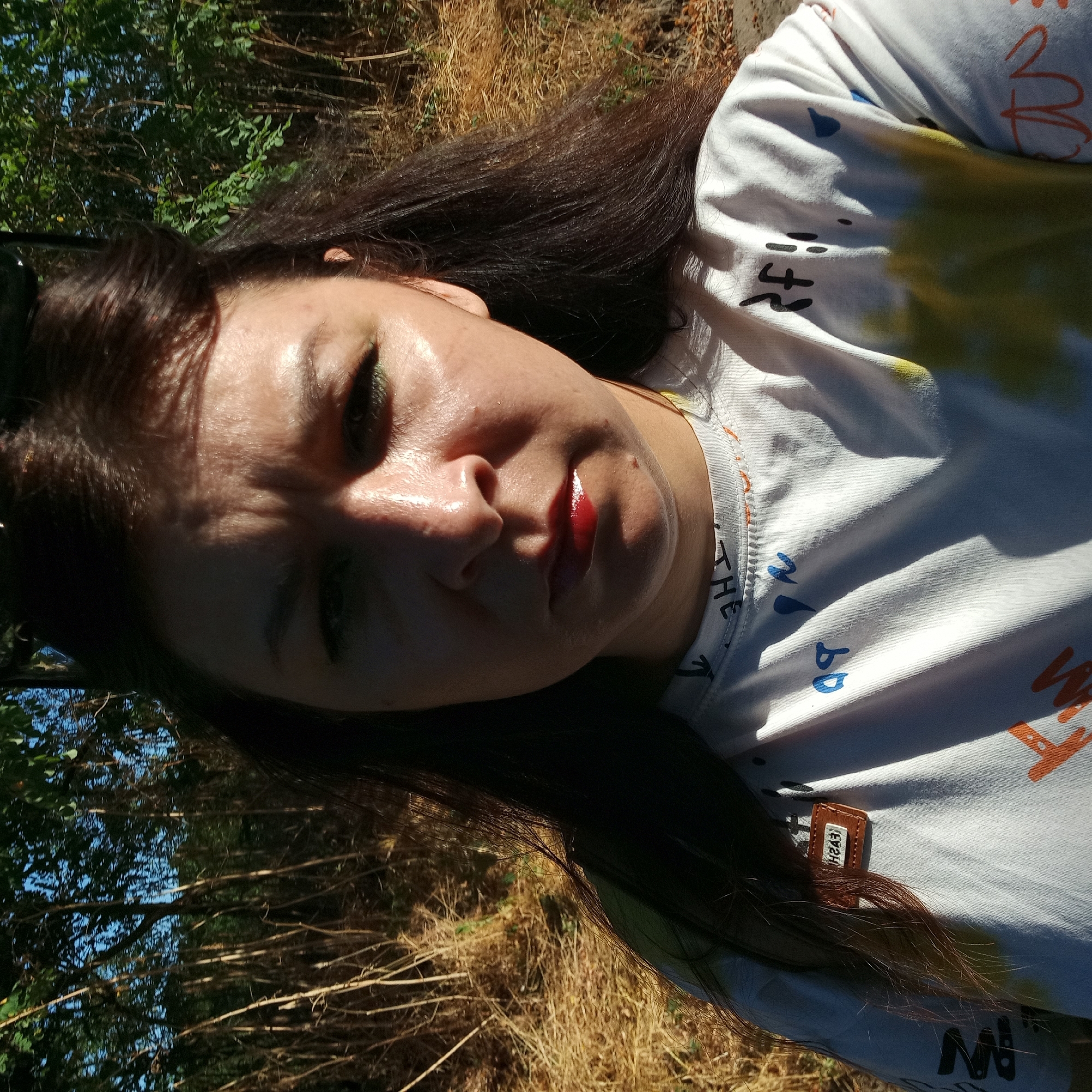 A medium shot is captured of a woman outdoors, facing the camera directly. Her face is illuminated by harsh sunlight, casting shadows. She has dark hair, pulled back from her face, and is wearing a white t-shirt with colorful graphic prints. A small brown tag with "FASH" printed on it in reverse is attached to the left side of her shirt. The background consists of dry grass and thin trees, suggesting an outdoor, possibly rural or park-like setting. There is no indication of the city of Klyntsi, Ukraine, in the image.