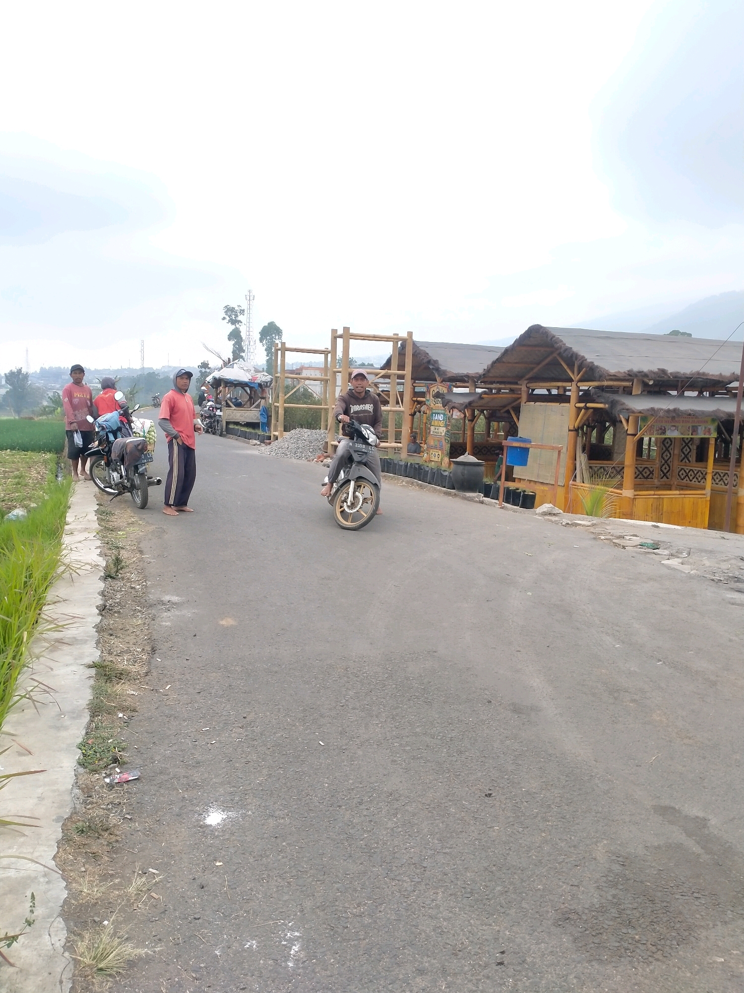 The image depicts a rural roadside scene in Batu, Indonesia, under an overcast sky. A paved road runs diagonally from the lower left to the upper right. On the left side of the road, a rice field with green stalks is visible, bordered by a concrete curb and a narrow strip of dirt and grass.
Several individuals are present. Three men are standing near a motorcycle parked on the left side of the road. One man, wearing a red shirt and dark pants, stands closest to the motorcycle. Another man, in a red long-sleeved shirt and dark pants, stands to his left, holding a dark object. A third man in a similar red shirt and dark pants is slightly behind them, wearing a dark cap. A fourth man, wearing a dark hooded sweatshirt and dark pants, is riding a motorcycle on the right side of the road, moving away from the viewer.
In the background, on the right side of the road, there is a structure with a thatched roof supported by bamboo poles. It appears to be a covered seating area or small establishment, with decorative bamboo elements and some signage. Several more motorcycles are parked near this structure. The landscape behind the structure suggests rolling hills and vegetation. The overall impression is a simple, functional roadside area in a village or rural setting.