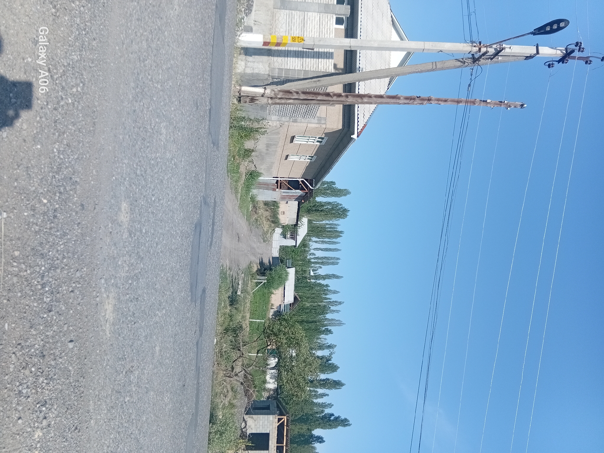 The image depicts a rural scene in the city of Kojo-Aryk, Kyrgyzstan, under a clear blue sky. In the foreground, a grey asphalt road leads into the distance. To the left, a concrete utility pole with a streetlamp and multiple wires is prominently featured. Adjacent to it, a wooden utility pole stands with wires running across the frame.
Further into the scene, a single-story house with a grey roof and white-trimmed windows is visible. Next to this house, a dirt road winds uphill. On the right side of the image, a partially constructed building with cinder block walls and exposed wooden roof beams is present. A mature tree with green foliage is situated in front of this construction.
A dense line of tall, slender trees forms a backdrop across the middle ground. The overall environment suggests a residential area with ongoing development and a natural, verdant landscape. The image is captured during daylight, with the sun casting shadows. A watermark indicating "Galaxy A06" is visible in the bottom left corner.