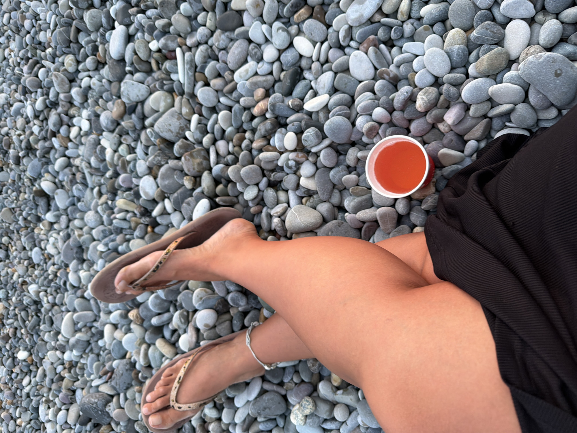 A person is relaxing on a pebble beach in Sochi, Russia, with their legs crossed and a cup of orange liquid beside them. Their feet are adorned with flip-flops and an anklet. The background is a dense arrangement of various-sized grey and white pebbles. The lighting suggests it is daytime.