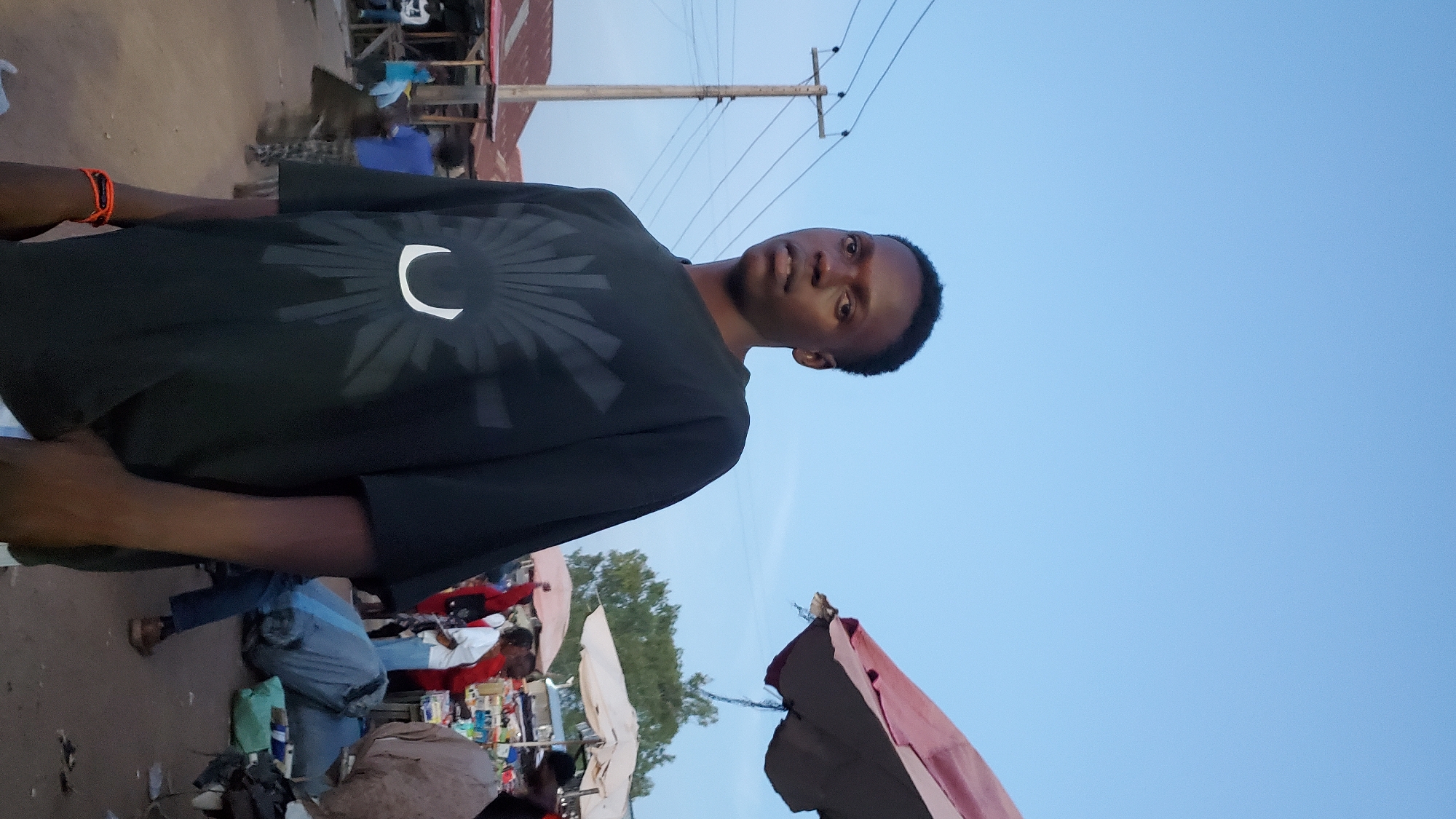 The image depicts a young Black male standing in what appears to be an outdoor market setting in Jalingo, Nigeria. He is wearing a black t-shirt with a white graphic design and has an orange bracelet on his left wrist. The man is looking towards the camera with a neutral expression. In the background, there are several market stalls with red and white awnings, various people browsing or working, and a wooden utility pole with electrical wires. The sky is a clear, pale blue, suggesting it is either early morning or late afternoon. The ground is dusty or unpaved. The overall scene suggests a typical day in a busy Nigerian market.
