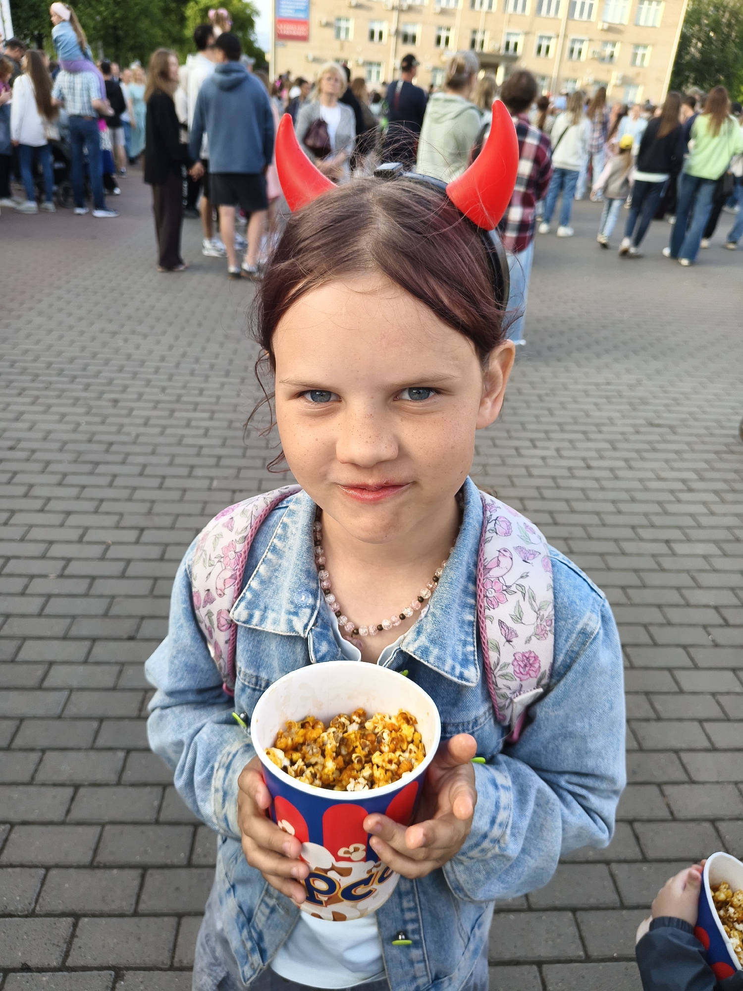 A young girl with red devil horns on her head and a denim jacket is holding a bucket of popcorn. She has brown hair and freckles. In the background, a crowd of people are walking on a paved area in front of a building. The image appears to have been taken outdoors.