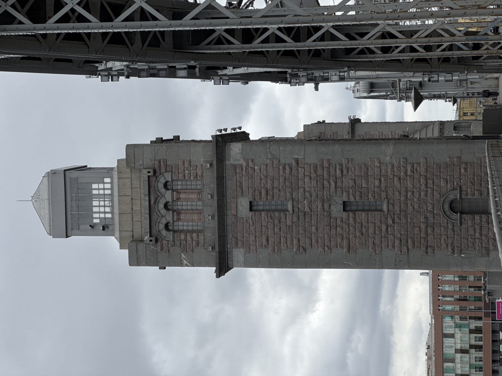 The image, oriented ninety degrees clockwise, captures a detailed view of architectural structures in Saint Petersburg, Russia, under an overcast sky. A prominent, tall, rectangular stone building or tower stands vertically in the mid-ground. Its facade is constructed from dark grey and reddish-brown rough-hewn stone blocks. The upper section features a lighter grey stone finish with a series of arched, multi-pane windows and decorative stone elements. Above these, simpler rectangular windows are set into the stone. The building culminates in a peaked, metallic roof section with a small spire and large, gridded windows.
Directly overhead, a massive metal bridge structure dominates the top of the frame. It comprises intricate grey trusses, girders, and riveted plates, characteristic of heavy-duty engineering. Numerous industrial-style lights are mounted on the underside of the bridge.
In the lower right background, a modern multi-story building with a brown and glass facade is partially visible, indicating an urban setting. The sky is uniformly grey and cloudy, suggesting an overcast day with flat, diffused lighting. No people or specific activities are discernible. A magenta sign with indistinct white text is partially visible on the modern building in the background.