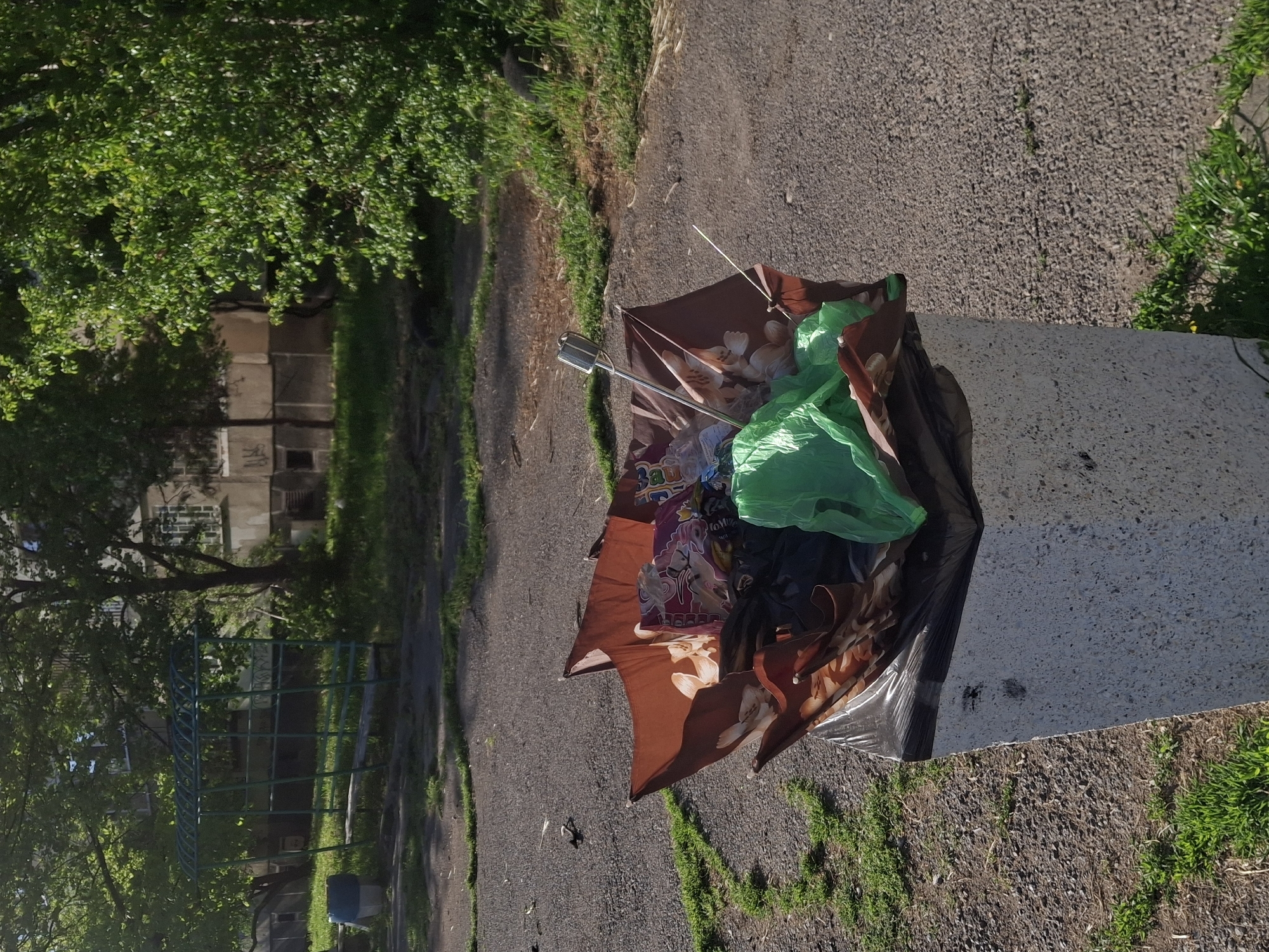 The image depicts a public space in Sofia, Bulgaria, near a playground and residential building. A concrete trash receptacle is overflowing with garbage, including a ripped shopping bag with images of women and flowers, several plastic bags including one green, other black and unknown colored bags. The bin appears overfull and the contents are spilling out. The surrounding environment includes asphalt pavement, patches of grass, and trees. In the background, there's a metal playground structure. The foliage suggests it's likely spring or summer. The buildings behind the trees appear to be residential.