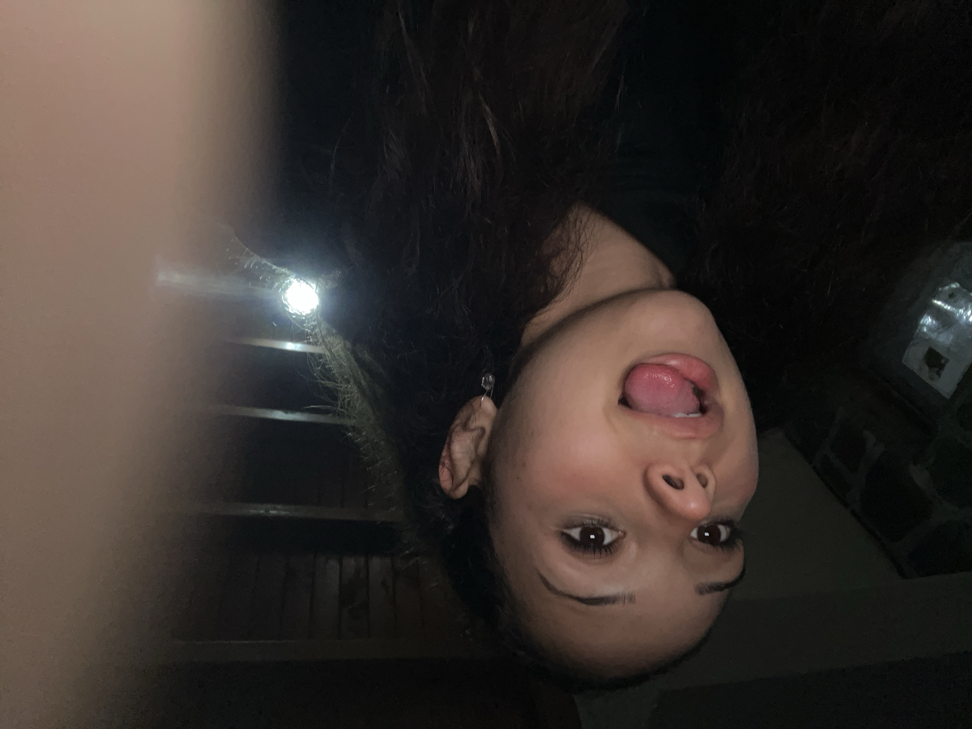 A young woman with long, dark, curly hair is taking a selfie in a dimly lit indoor setting. She has her tongue sticking out and her mouth open, with a playful expression. She is wearing a dark hoodie. There is a blurry light source in the background, and some indistinct shapes that could be furniture or architectural features. The image is a close-up and the lighting is very low, creating a dark atmosphere. There is no visible indication of the city of Zlatograd, Bulgaria within the image itself.