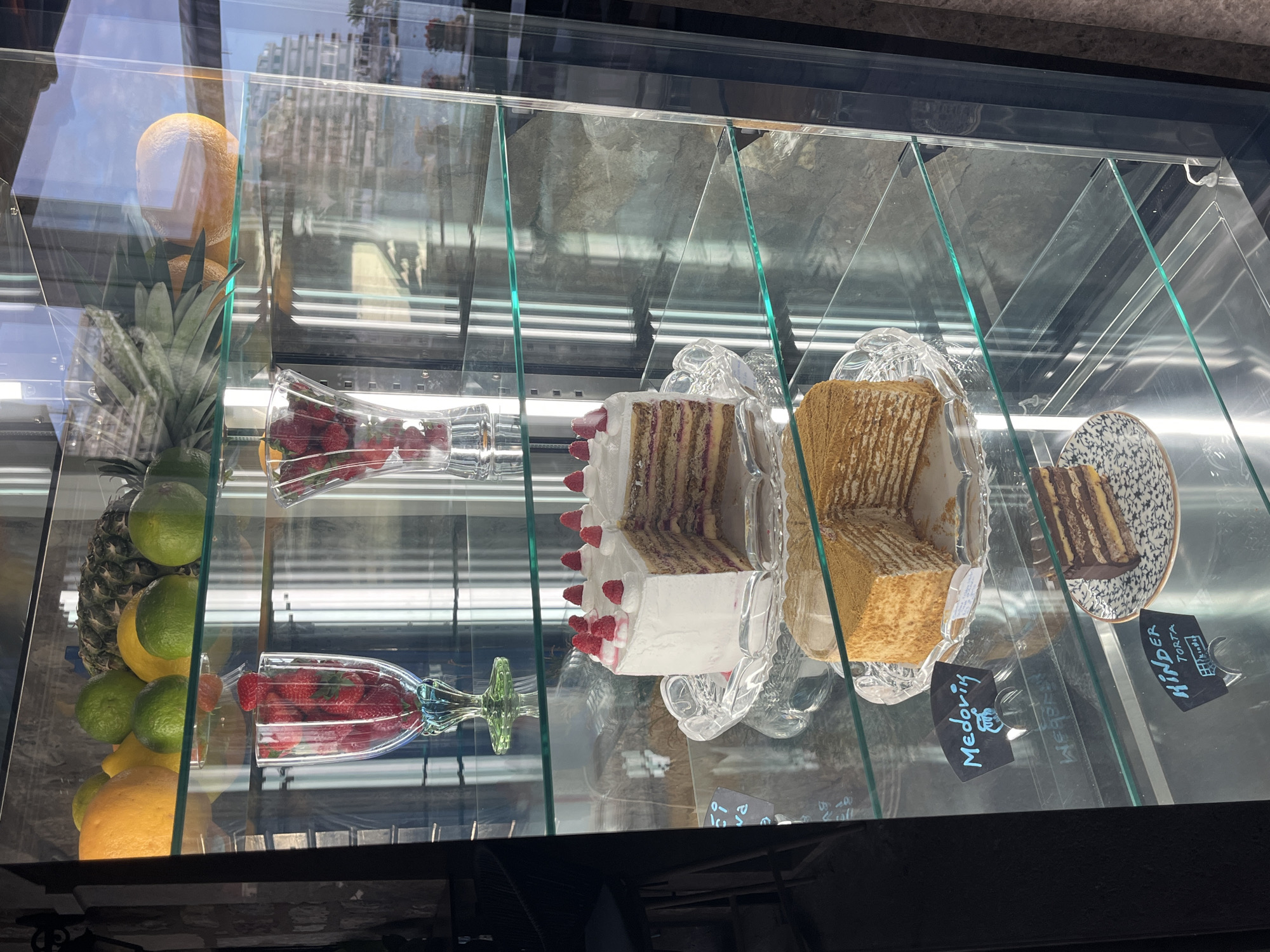 The image shows the interior of a refrigerated display case containing a selection of desserts, likely within a commercial establishment. The case is located in Boreti, Montenegro.
On the left side of the case are various fruits, including oranges, limes, and a pineapple. Two glass vases hold strawberries.
Three different types of cake are displayed on decorative glass plates. From left to right, they appear to be a raspberry and cream cake, a honey cake ("Medovik" according to the small sign), and another cake labeled "Kinder Torta."
The case appears to be clean and well-lit. The reflective glass of the display case causes some distortion and shows reflections of the surrounding environment.