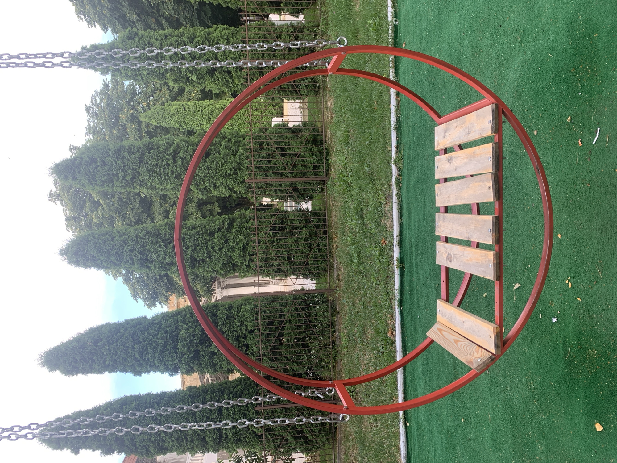 This is a medium-angle, eye-level, outdoor shot of a red, circular swing with a wooden seat, likely located in a park or recreational area in Dondușeni, Moldova. The swing is suspended by four metal chains, two visible on each side. The swing's circular frame is made of red metal tubing, and its seat is composed of several wooden planks. The ground beneath the swing is covered in green artificial turf, with some patches of grass and scattered leaves. In the background, a series of tall, dark green evergreen trees are visible, planted behind a dark metal fence. Behind the trees, a light-colored building with architectural columns can be seen. The sky above is overcast. There are no people present in the image.