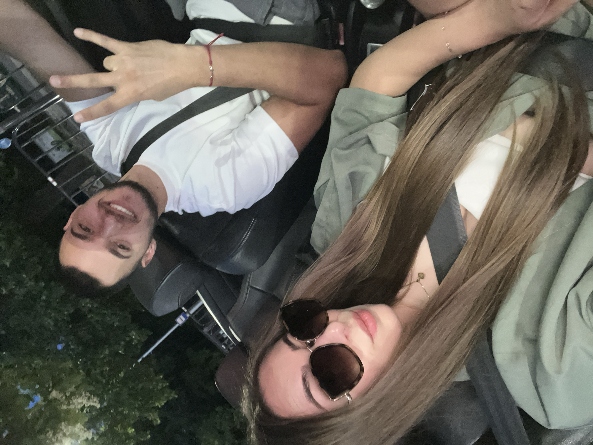 In this image, a young man and woman are seated in a vehicle. The woman, in the foreground, wears large, tortoiseshell sunglasses and a green jacket, her long brown hair flowing over her shoulders. A delicate gold necklace is visible around her neck. The man, positioned behind her, is smiling and making a "rock on" hand gesture. He is wearing a white t-shirt and a red string bracelet. Both are wearing seatbelts, indicating they are likely in motion. The background shows foliage and what appears to be a metal railing, suggesting an outdoor setting, possibly in Plovdiv, Bulgaria, given the context of the prompt. The lighting is dim, suggesting it is evening or nighttime. The scene is captured from a close-up perspective, focusing on the couple.