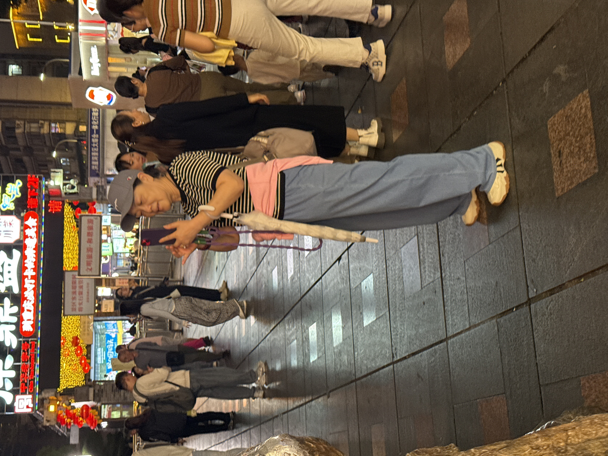 Here is a description of the image:
This image captures a crowded outdoor scene, likely in the Yuzhong District of China. The composition is taken at eye level, showing a sidewalk paved with rectangular gray stone tiles. A group of people are present, primarily walking or standing, and some seem to be looking at or interacting with their phones.
In the foreground, an older woman lies prone on the sidewalk, presumably for a photograph, wearing a striped shirt, light blue pants, white sneakers, and a baseball cap. A folded umbrella lies beside her. Several other individuals can be seen behind her, standing and looking in the same direction as the woman on the ground.
The background features brightly lit storefronts, with numerous signs in Chinese characters, some illuminated with neon lights and colorful decorations. The lighting indicates that it is nighttime. The overall scene depicts a lively urban setting, possibly a tourist area or shopping district.