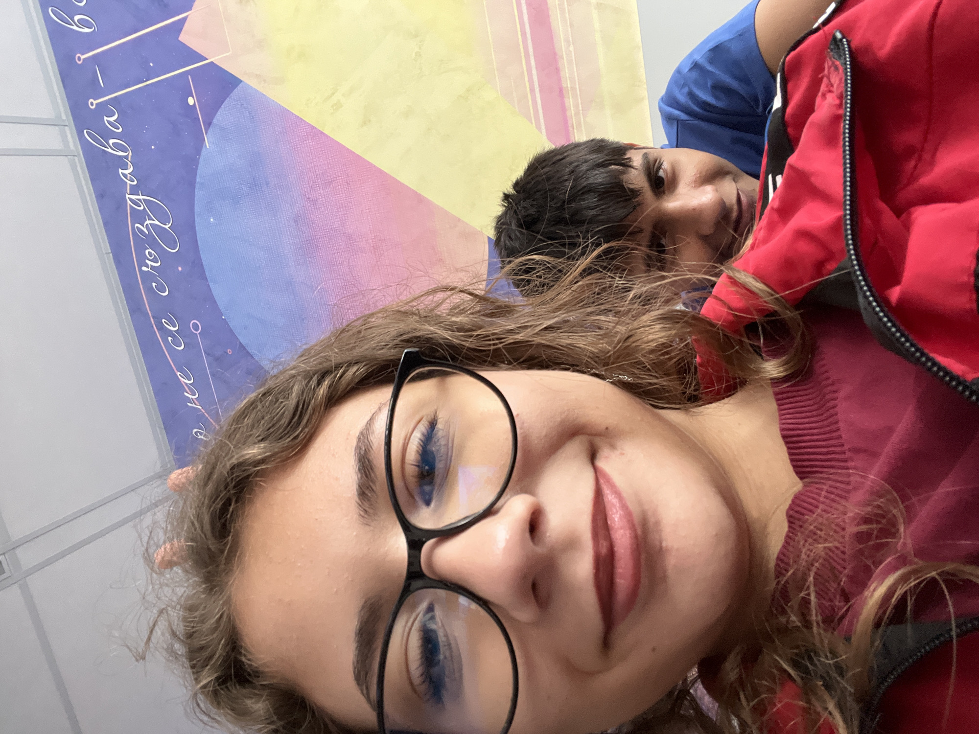 A selfie captures two young individuals indoors, likely in Plovdiv, Bulgaria. In the foreground, a female with long, wavy brown hair and black-rimmed glasses smiles towards the camera. Her blue eyes are visible, reflecting light from the lenses. She wears a burgundy ribbed top. Behind her and to the right, a male with dark hair looks towards the camera, wearing a blue shirt and a red, zippered jacket.
The background features a colorful mural on a wall, depicting abstract geometric shapes and celestial elements in shades of purple, blue, yellow, and pink. White lines and small circles are also part of the design. Vertically oriented Cyrillic text is visible on the purple section of the mural, including the phrase "се създава" (is created). The scene conveys a casual and friendly atmosphere.