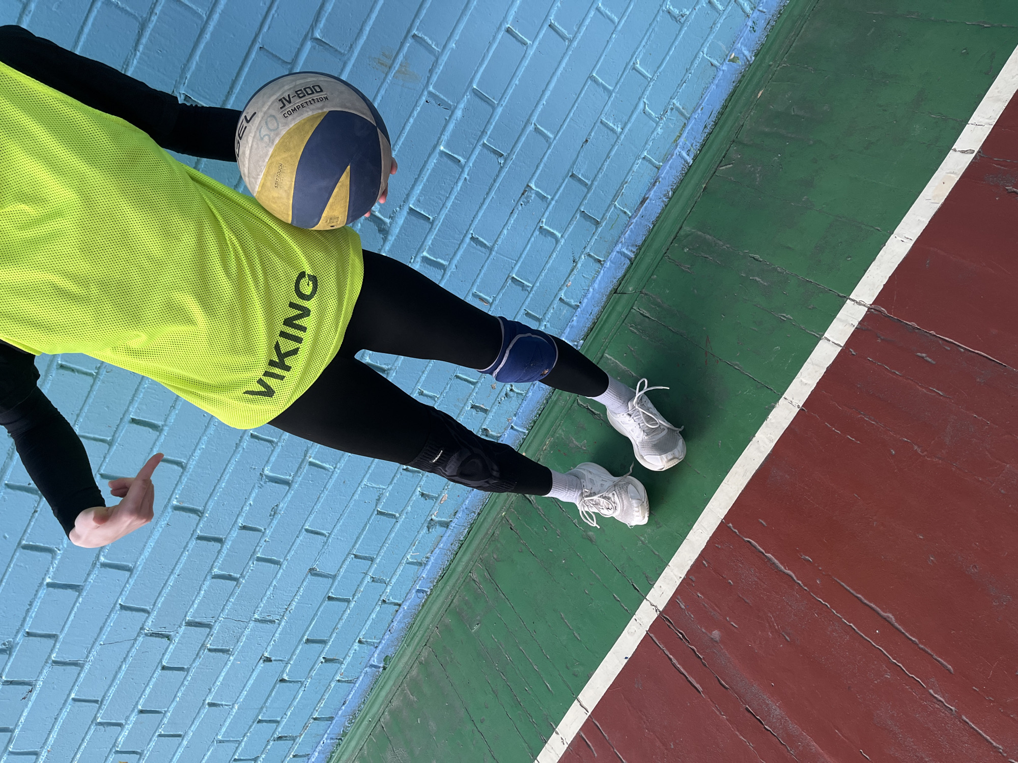 A high-angle, tilted photograph captures the lower body of a person holding a volleyball within an indoor sports facility in Saint Petersburg, Russia. The individual is dressed in black long-sleeved athletic wear and black leggings. Over the black top, they wear a vibrant neon yellow mesh vest with the word "VIKING" printed vertically in black letters. Visible on their knees are pads, one blue on the left leg and a black one on the right. They are wearing white athletic shoes with white laces and white socks.
The volleyball, held in the person's hands, is multicolored in white, yellow, and blue panels. Text visible on the ball includes "JV-800", "COMPETITION", and "SOFT TOUCH," with the brand "MIKASA" partially visible and inverted.
The background consists of a bright blue painted brick wall to the left. The floor is made of worn wooden planks, painted in sections of green and reddish-brown, divided by a distinct white line. This arrangement strongly suggests a sports court. The bright lighting indicates daytime. The scene depicts a moment likely during a volleyball practice or game.