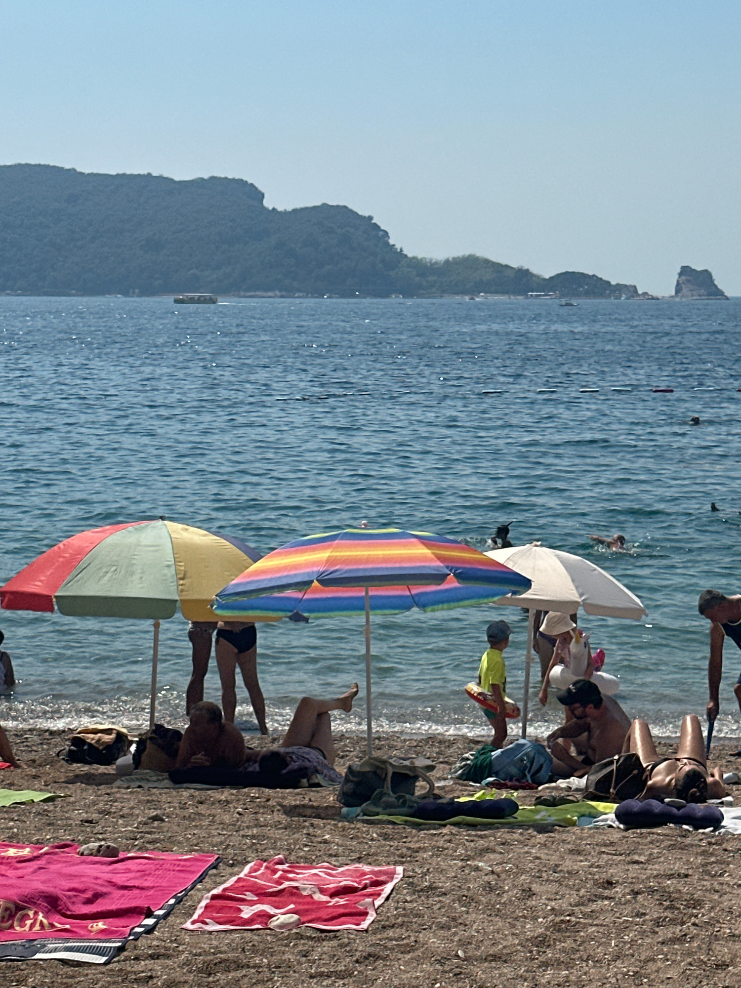 The image depicts a sunny day at a beach, likely in Budva, Montenegro, with the Adriatic Sea and a mountainous coastline in the background. Several beach umbrellas, including a multi-colored striped one and a red and green one, provide shade for beachgoers. People are seen sunbathing on towels and in the water, swimming and enjoying the sea. In the foreground, various beach towels and bags are laid out on the sand. The water is clear blue, and in the distance, a small boat is visible near the shore. The scene captures a typical summer day at a popular coastal destination.