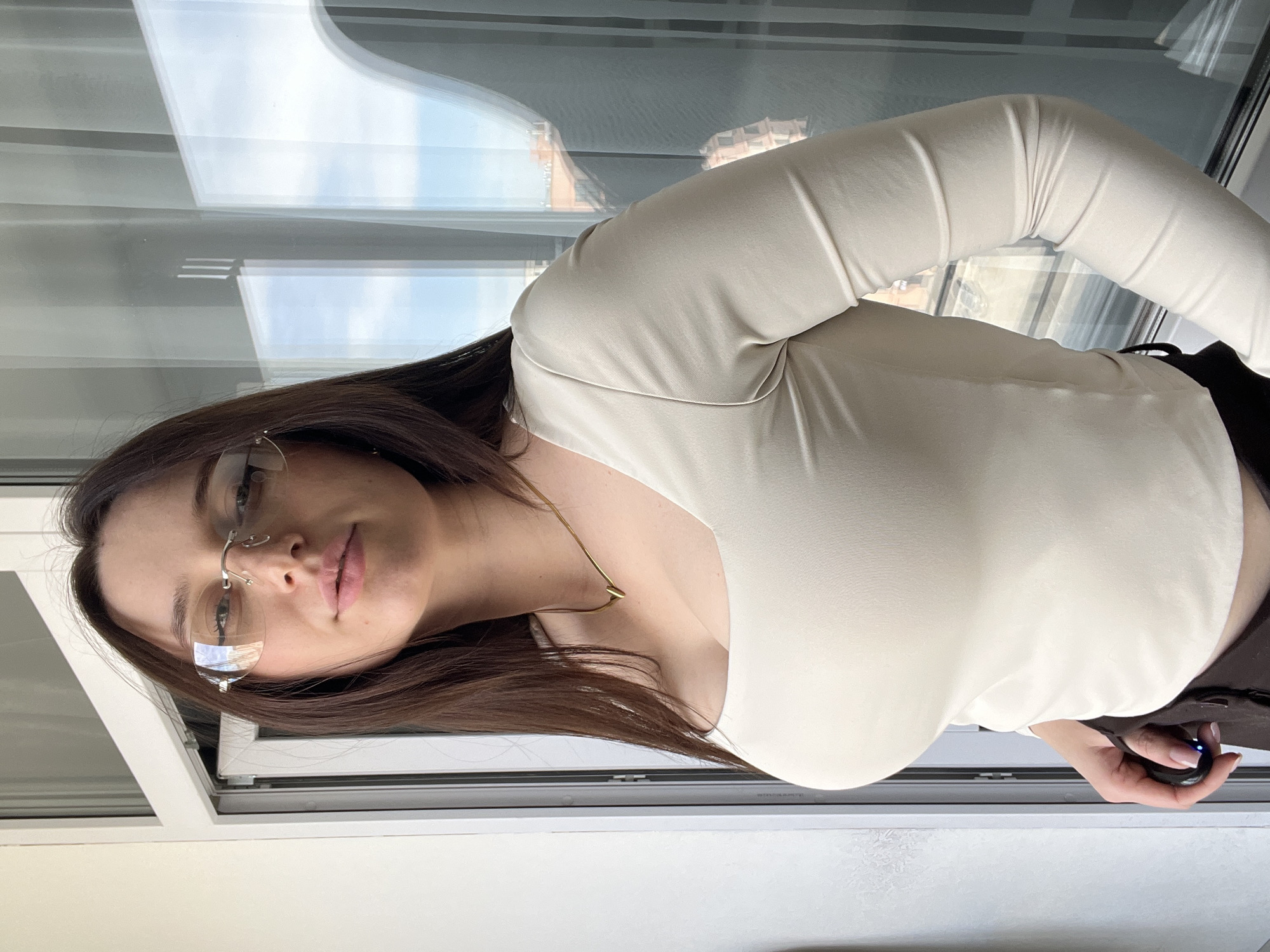 A woman with long brown hair and glasses is standing near a window, possibly inside an apartment or building in Tyumen, Russia. She is wearing a light-colored, long-sleeved top and a gold necklace. She is holding a black object, possibly a remote or electronic device, in her right hand. The window provides a view of the city. The scene appears to be indoors.