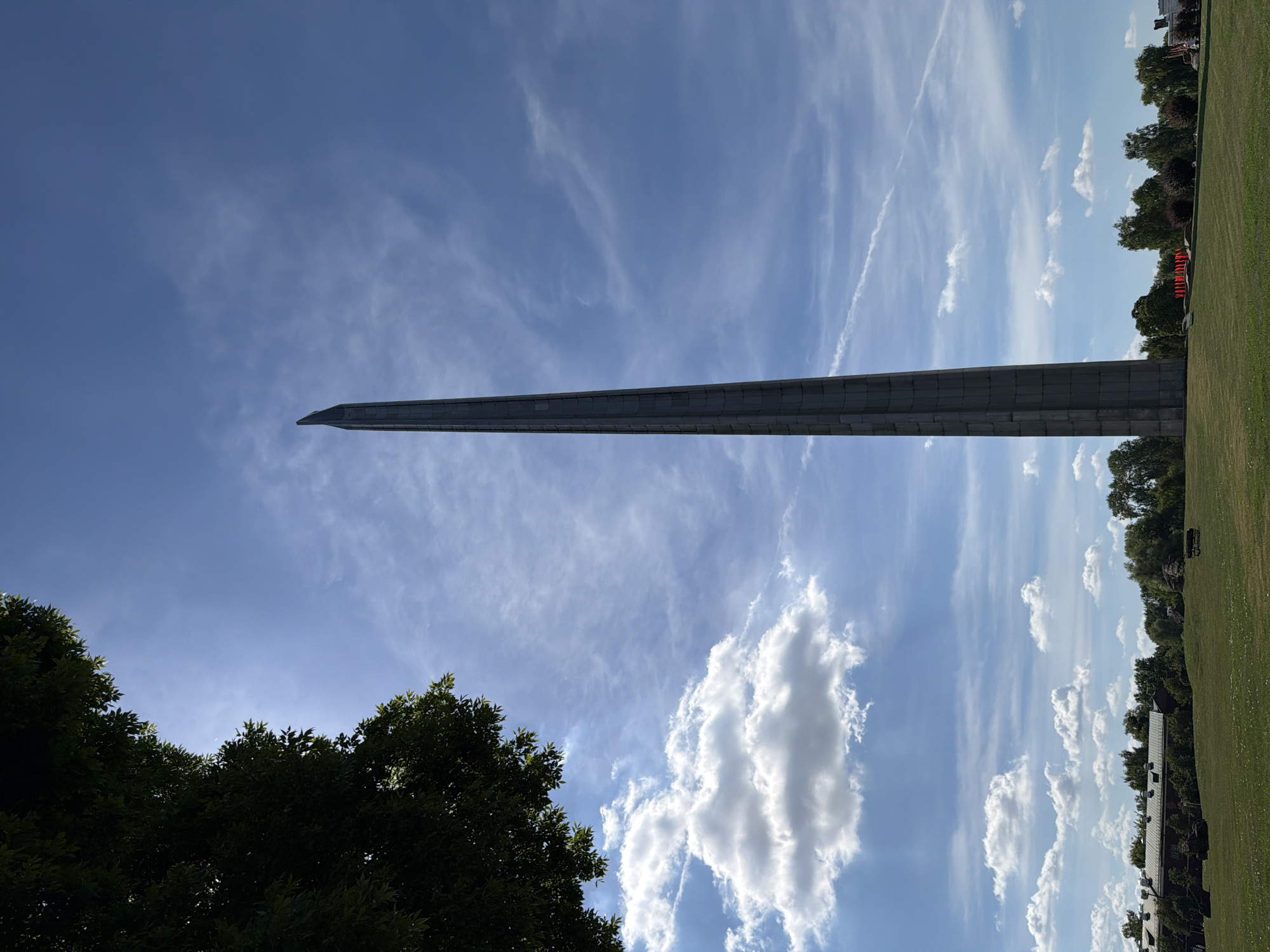 This media file depicts an outdoor scene on a clear, sunny day. The dominant subject is a tall, slender, gray obelisk or monument that stretches diagonally across the frame from the lower left towards the upper right. Its surface appears to be constructed of segmented, rectangular blocks.
In the foreground, on the left side of the image, the dark green foliage of trees frames the lower portion. To the right of the obelisk, a manicured green lawn slopes gently upwards, bordered by a line of trees and some buildings in the distance. Wispy white clouds are scattered across the bright blue sky, with some larger, fluffier clouds also present. The overall impression is one of a monument situated in a park or memorial ground. The lighting suggests it is daytime, likely late morning or afternoon, given the bright sun. There is no visible text in the image.