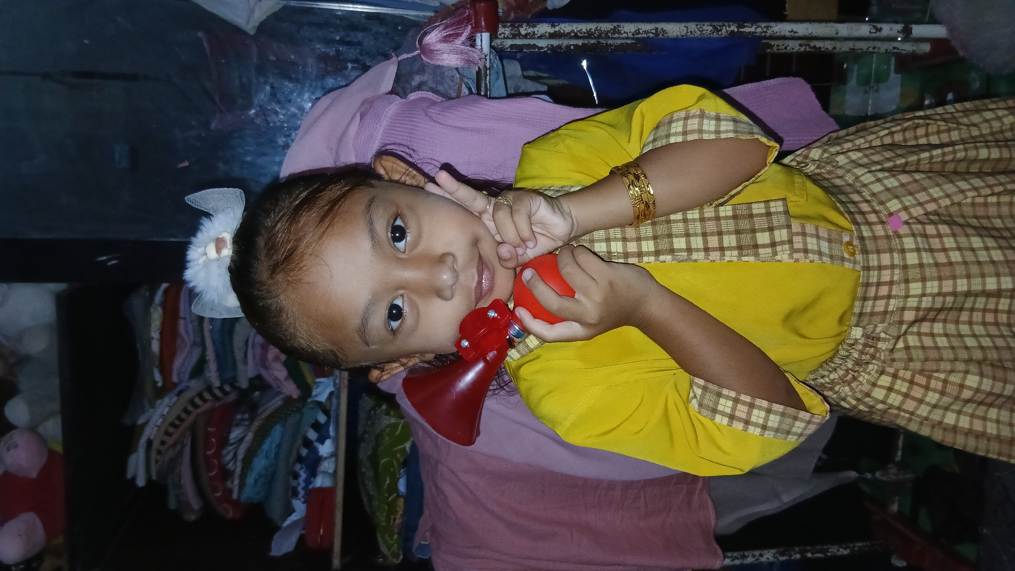 A young girl, likely of Southeast Asian descent, is positioned centrally in the frame, holding a red novelty horn to her mouth with her right hand. She is wearing a bright yellow shirt with plaid trim and a matching plaid skirt. Her left hand is raised, displaying two fingers in a peace sign. She has a gold bracelet on her right wrist and two rings on her left index finger. A white hair accessory with a small animal charm adorns her brown hair, which is pulled back. The background consists of shelves filled with folded textiles and fabric, suggesting an indoor setting, possibly a home or storage area, in Keudejungkagajah, Indonesia. The lighting is dim, with the subject being the primary light source.