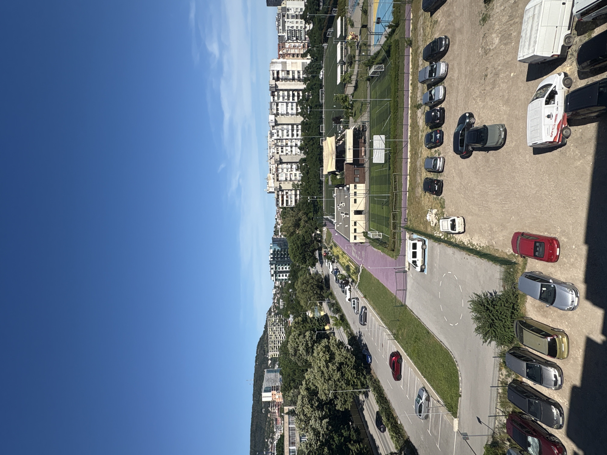 This media file shows an aerial view of a city scene. The main subjects are buildings, a parking lot with several cars and vans, a green sports field, and a clear blue sky with some wispy clouds. The scene is set in an urban environment, with a mix of modern and older buildings visible on a hillside.
The image appears to be taken on a sunny day, likely during the daytime, as indicated by the bright lighting and the clear sky. The orientation of the image suggests it was taken from a high vantage point, possibly a tall building.
Notable details include a variety of vehicles: sedans, vans, and what appear to be SUVs, parked in rows. One white van has "КАРТИНКИ НОВЫХ" visible on its side. Another white van, possibly a camper or recreational vehicle, is parked near a row of cars. A red car is parked in a designated spot, and several other cars are scattered throughout the parking area. The sports field has markings and what look like goalposts. Trees and greenery are interspersed with the buildings and urban infrastructure.
The overall impression is one of a typical urban landscape during daylight hours. There are no people prominently visible in the image, and no specific event appears to be taking place, other than the everyday activities of a city.
