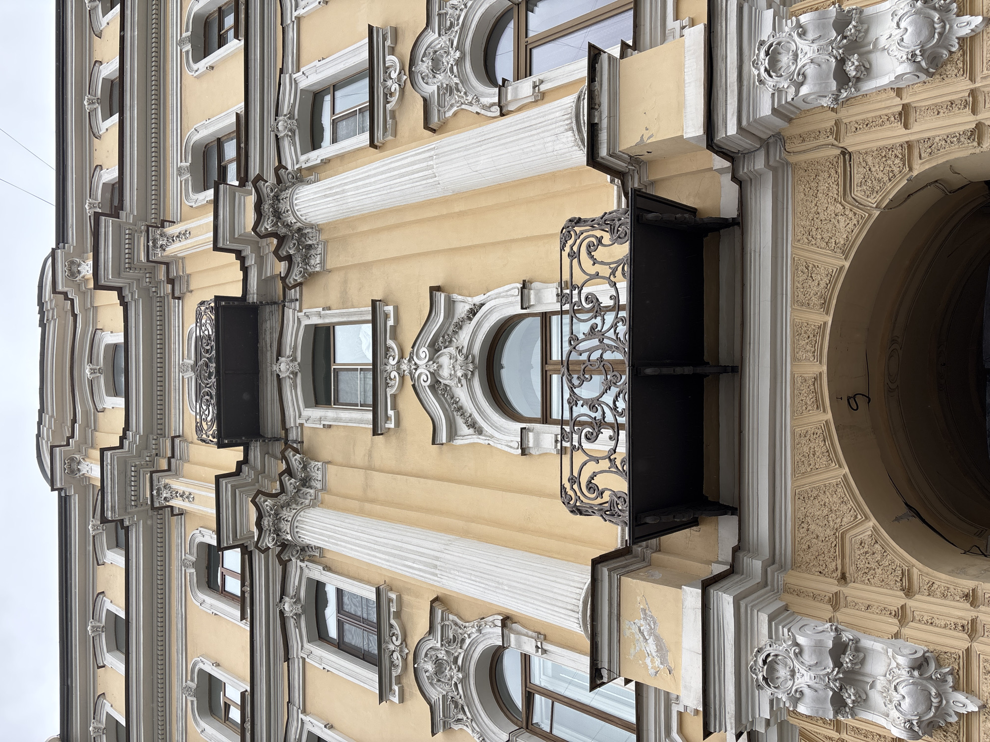 This image depicts a section of an ornately decorated building facade in Saint Petersburg, Russia. The building is multi-storied with a yellow exterior and elaborate white architectural details and moldings. There are a series of windows, some rectangular and some arched, each framed with decorative elements. Ornate iron balconies are attached to the building, offering decorative detail as well as access. The architectural style features classical influences, with columns, pilasters, and decorative carvings. The overall impression is one of grandeur and historical significance, typical of the architecture found in Saint Petersburg.
