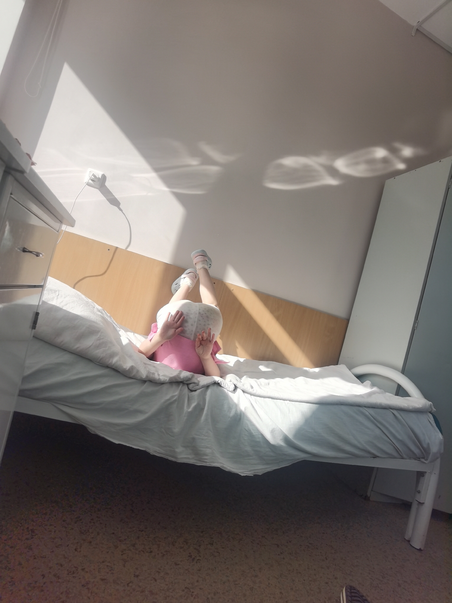 A child is lying on their back on a hospital bed. The child is wearing a pink shirt, white diaper, and light blue shoes. The bed has a white metal frame and a light blue sheet. A cabinet is on the left side. The wall is beige and white. The location is likely a hospital room in Perm, Russia. Light from a window casts shadows on the wall and ceiling.