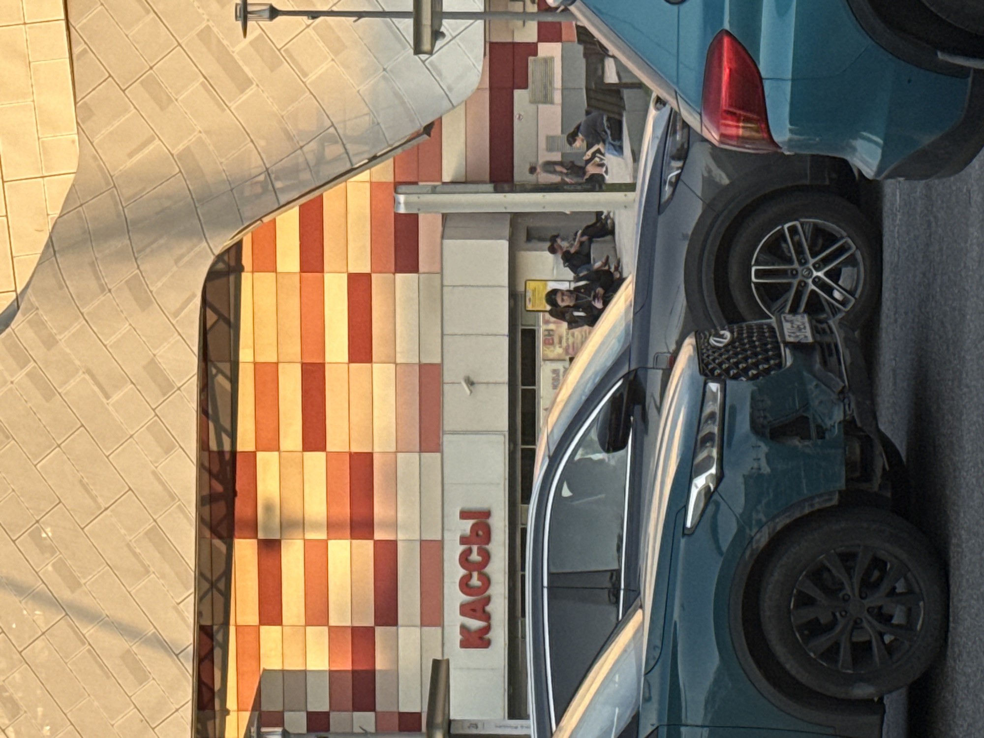 A partially obscured view of a commercial or public building with a checkered exterior wall and the Russian word "KACCDI" (KASSY) vertically aligned, likely indicating "cash desks" or "checkouts," is framed by the front and rear portions of two vehicles parked in the foreground. The building facade features horizontal bands of tiles in alternating shades of beige, orange, and red. People are observed seated near the building's entrance. A textured, curved wall is visible to the left of the checkered facade, suggesting architectural complexity. The urban setting, along with the Russian word, points towards Moscow, Russia, as the potential location.