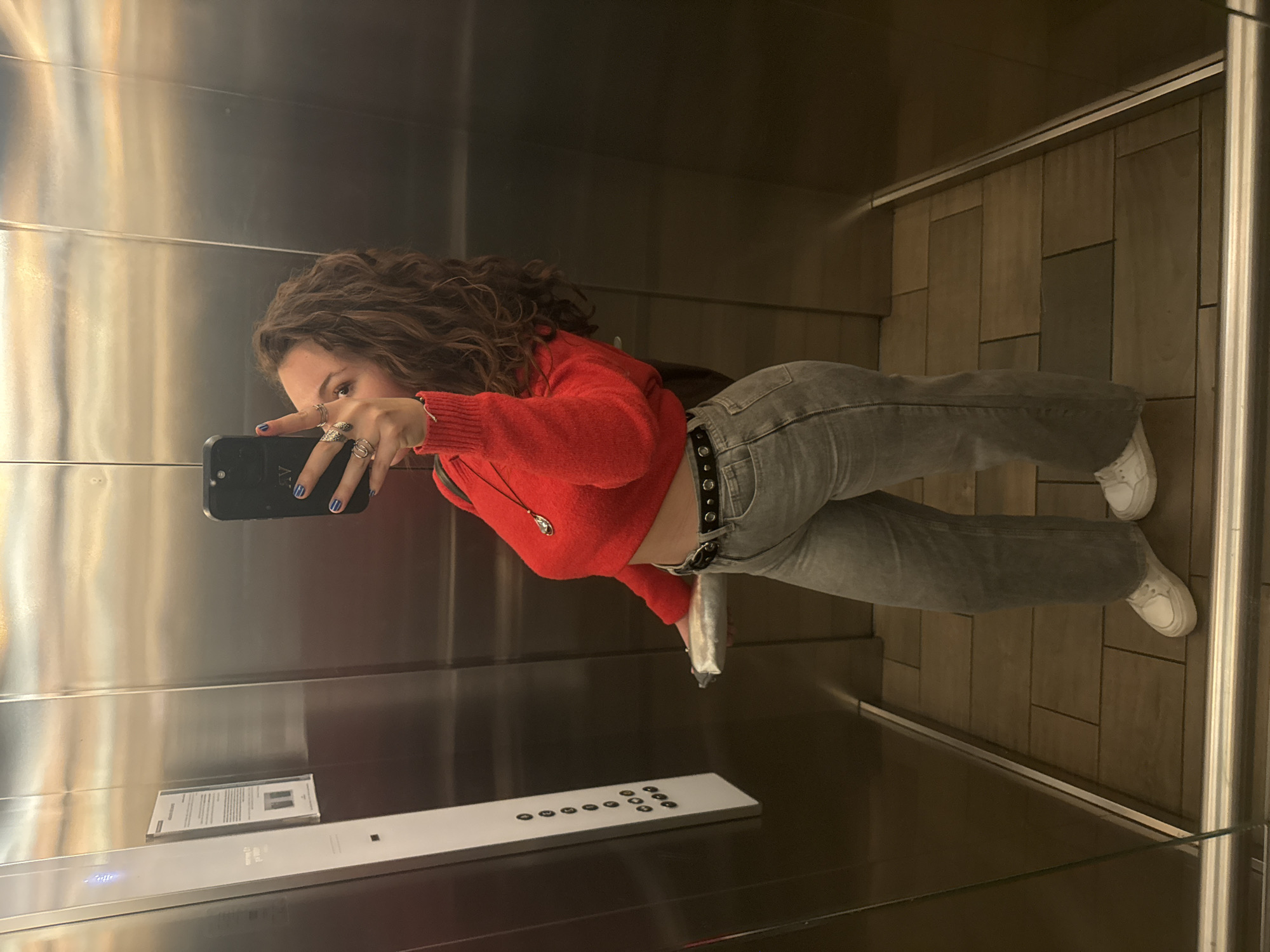 A young woman is taking a selfie in an elevator. She has curly brown hair and is wearing a red cropped sweater, gray jeans with a black studded belt, and white sneakers. She is holding a black phone in her left hand, which has blue nail polish and multiple rings. The elevator has metal walls and a control panel with circular buttons. The floor is tiled in a brown and gray pattern. A sign with text is visible on the elevator wall. The location is potentially in Mexico City, Mexico.