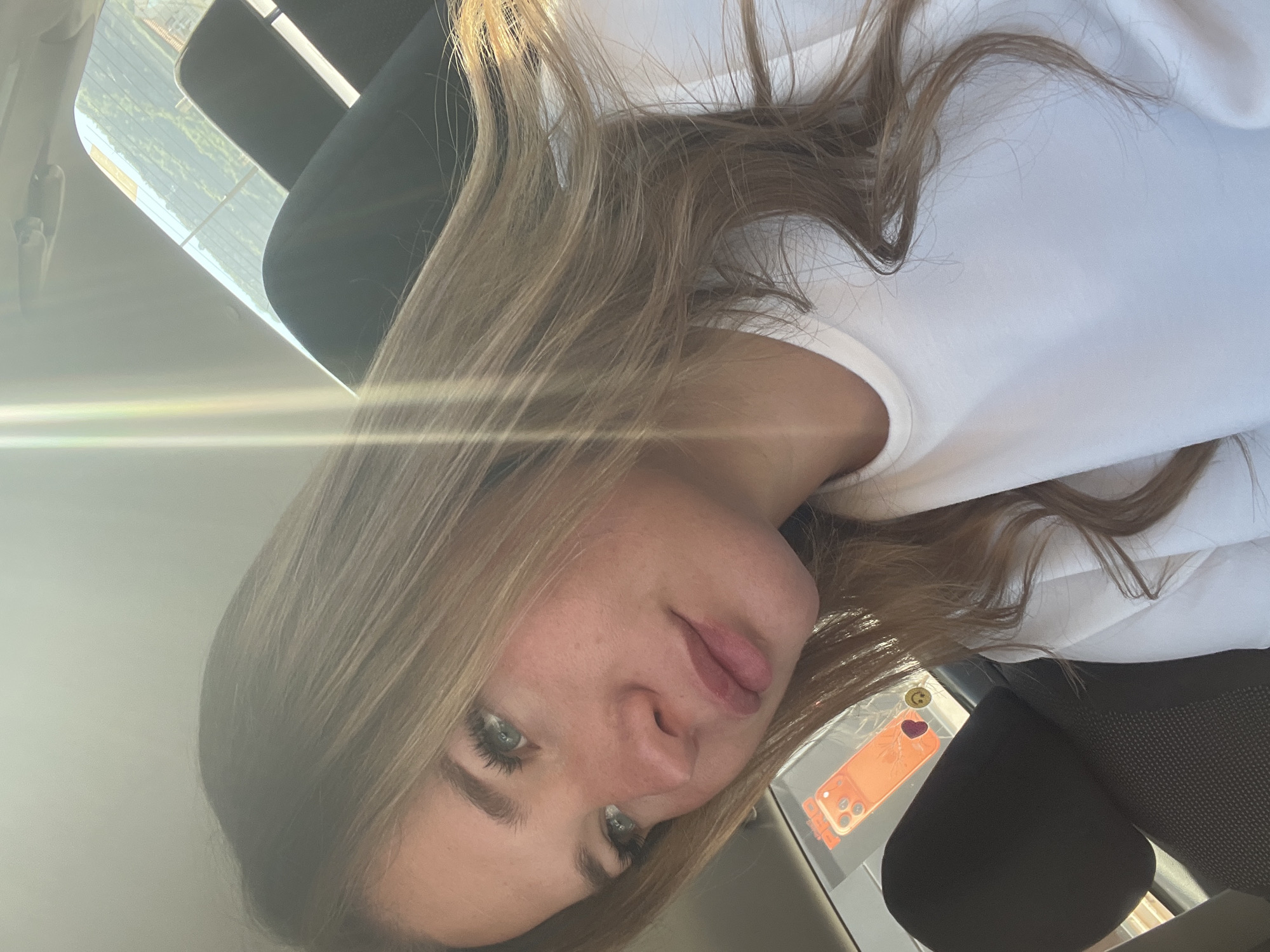 A young, fair-skinned woman with blonde, shoulder-length hair and green eyes is seated in what appears to be a car. She's wearing a white t-shirt. The car's interior is visible, including a black headrest and possibly the dashboard. A mobile phone with an orange case and a heart sticker attached is visible in the lower right corner. Outside the car, there is the suggestion of an urban landscape through the car window. There is a bright horizontal glare in the upper left of the frame, possibly from the sun. There is not enough information to place this image in Budva, Montenegro.