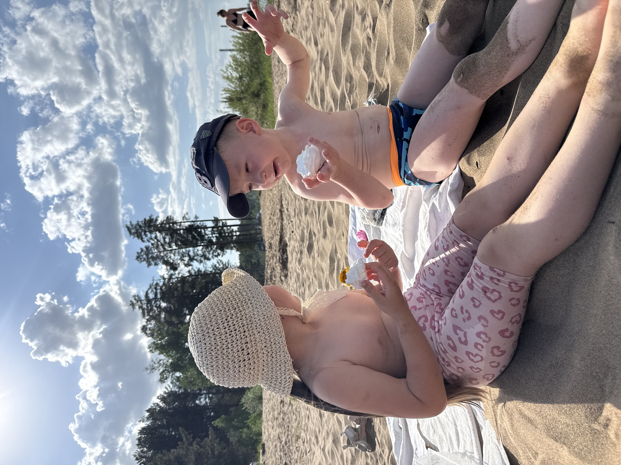 The photograph captures two children on a sandy beach, potentially in or near the city of Chaikovsky, Russia. The boy, wearing a dark baseball cap and blue swimming trunks with orange accents, is holding a ball of what appears to be white foam or cream. His arm is raised in the air. The girl, wearing a light-colored crocheted hat and pink patterned leggings, is also holding a similar object. They are positioned on a white towel laid out on the sand. The background shows trees and a sky with scattered clouds. The environment seems to be a sunny day at a beach.