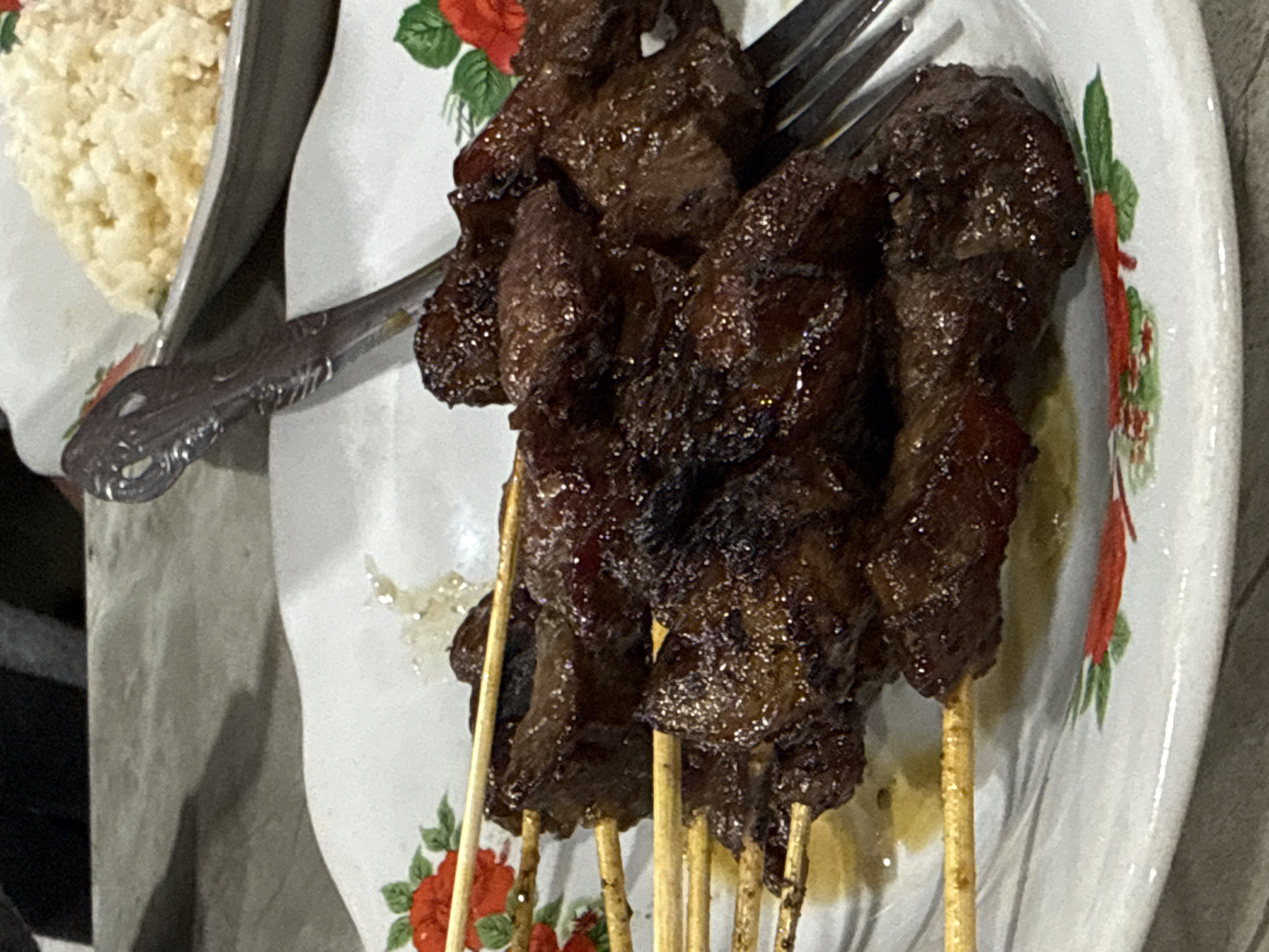 The image shows a plate of dark brown, glossy meat skewers, likely satay, served with a portion of white rice. The skewers are arranged on a white plate decorated with red rose and green leaf patterns. A fork rests on the plate near the skewers. The setting appears to be a food serving, possibly in a restaurant or home, with a partially visible blurred background. The city of Payameuneng, Indonesia, is not discernible from the image, therefore I can't include it.
