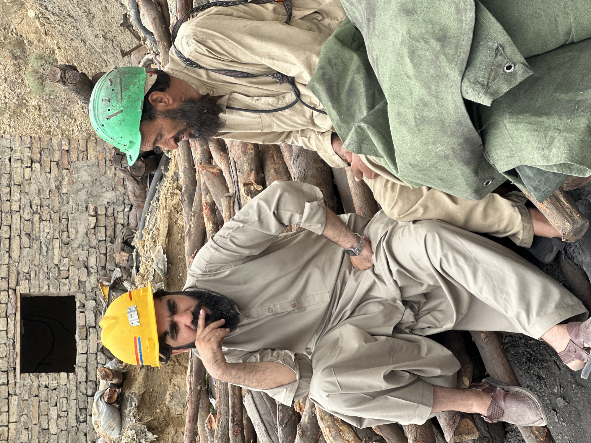 Two men wearing work clothes and hard hats are seated on a pile of logs in what appears to be a construction or industrial area. The man on the left is wearing a yellow hard hat and a light-colored shalwar kameez, with his hand on his chin. The man on the right is wearing a green hard hat and a similar light-colored outfit, with a beard and a strap over his shoulder. In the background, there is a brick wall with a dark opening, and more logs and debris. The ground is covered in dirt and smaller pieces of wood. The scene suggests a break or pause in work activity in an outdoor environment that may be located in the city of Quetta, Pakistan, a region known for its mining and construction activities.