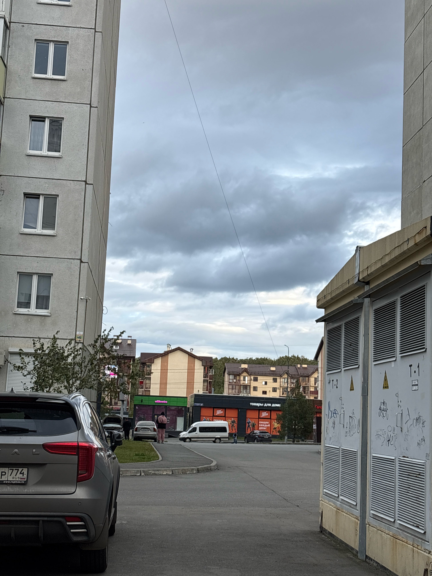 The image captures a street scene in Zapadny, Russia, under an overcast sky. On the left, a tall, grey, concrete apartment building with rows of windows dominates the foreground. To the right, a large, grey electrical transformer box with ventilation louvers and markings like "T-1" and "T-2" stands prominently.
In the mid-ground, across the asphalt road, a row of buildings is visible. One building features an orange and beige striped facade, while another has a dark facade with illuminated signs, including one with Russian text that appears to say "ТОВАРЫ ДЛЯ ДОМА" (Goods for the Home). Several vehicles are parked or driving on the street, including a grey SUV in the immediate foreground on the left, a white van, and several other cars. A few individuals are present on the sidewalk, one of whom appears to be walking away from the camera. Trees and other foliage are scattered throughout the scene, and utility poles and wires are also visible.
