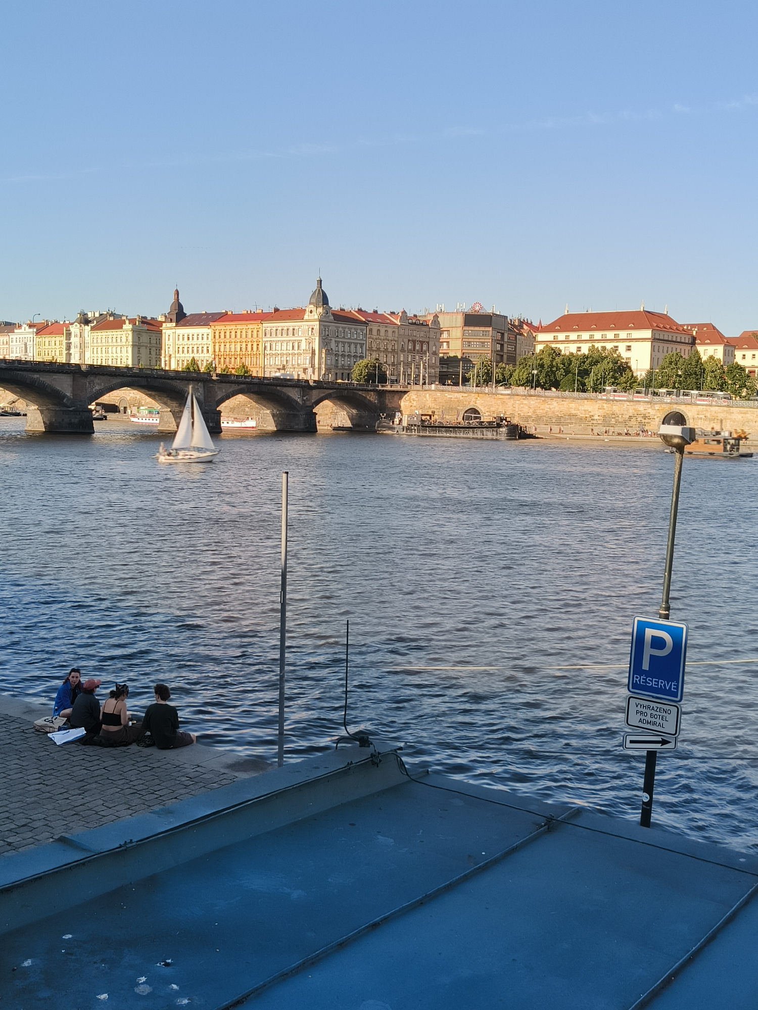 The image captures a scenic view of the Vltava River in Prague, Czechia, during daylight hours. In the background, a bridge with multiple arches spans the river, connecting to a cityscape characterized by a row of historic buildings with red-tiled roofs and light-colored facades. A white sailboat navigates the rippling water of the river.
In the foreground, on the paved embankment to the left, four individuals are seated. They appear to be casually conversing or relaxing by the water. A metal pole with a sign indicating "P RÉSERVÉ" and further text below is visible to the right, suggesting a reserved parking area. The river's surface is textured with gentle waves, reflecting the light and the surrounding environment. A section of a dark, metallic structure occupies the bottom of the frame, possibly a railing or a part of a building overlooking the river. The sky is clear and blue, indicating pleasant weather.