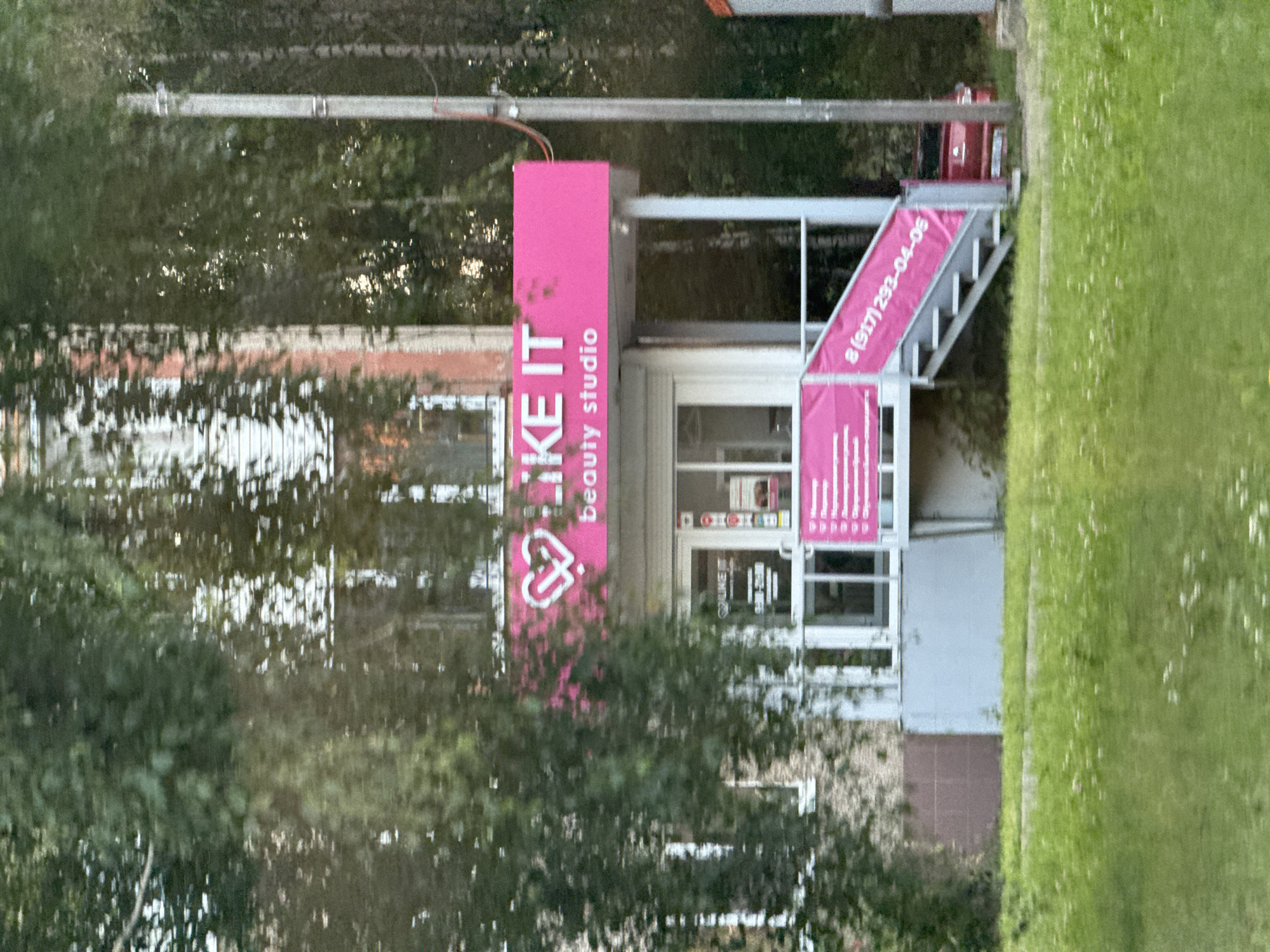 The image shows the exterior of a beauty studio named "LIKE IT" in Izhevsk, Russia. The studio is located on the ground floor of a building, with a pink awning above the entrance displaying the name "LIKE IT" and "beauty studio" in white lettering. Below the awning, there is a glass-paneled door and windows. A pink banner with text in Cyrillic is visible on the stairs leading to the entrance. The building appears to be brick and has windows on the upper floors. Trees and foliage surround the studio, and a red car is parked to the right of the entrance. The area in front of the building is covered in green grass with small white flowers.