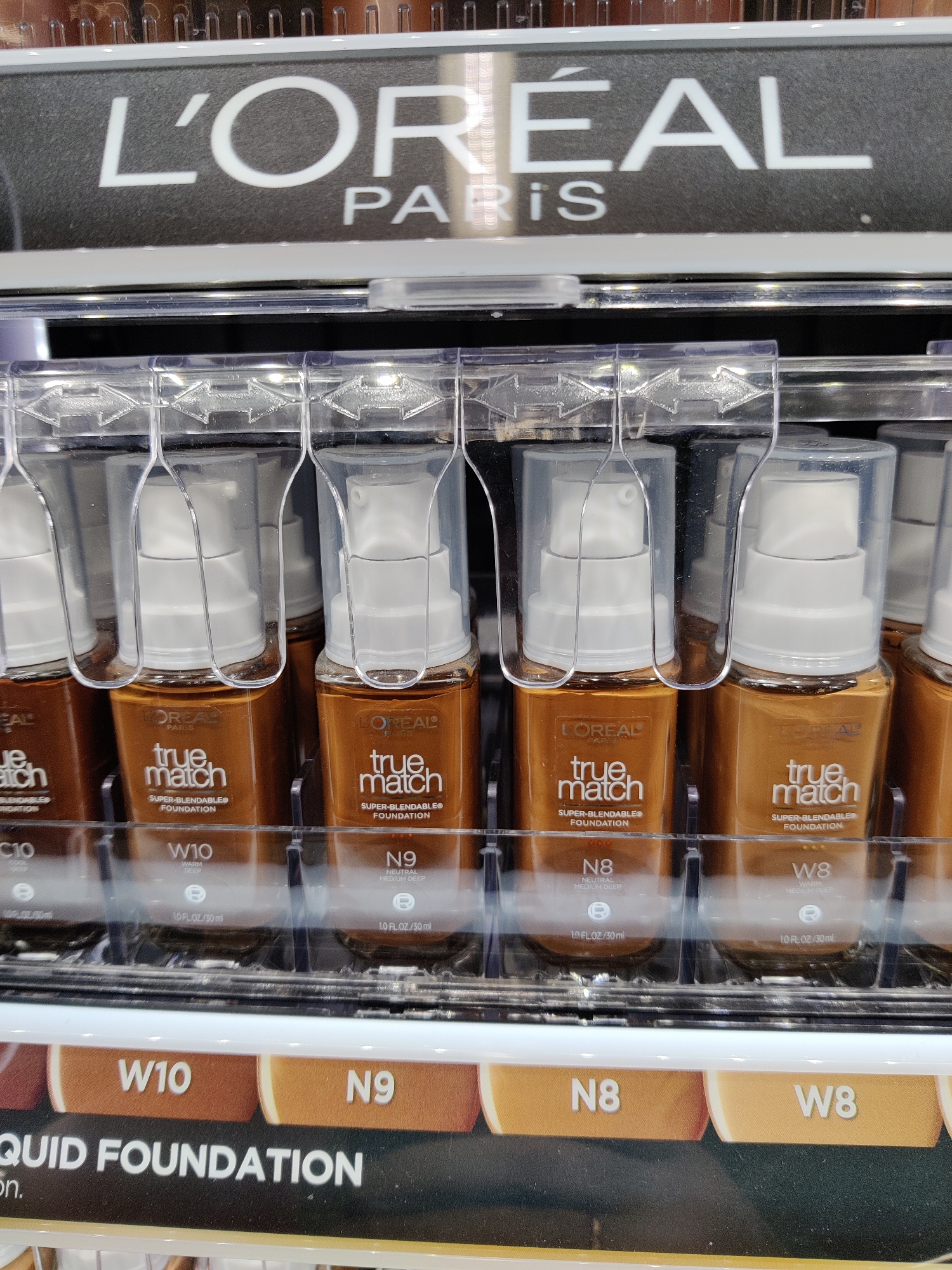 The image displays a retail shelf filled with L'Oréal True Match liquid foundation bottles. The bottles are arranged in rows, each with a clear plastic pump dispenser. The brand name "L'ORÉAL PARIS" is prominently displayed on a header above the products. Below the bottles, color swatches labeled with shade numbers like "W10," "N9," "N8," and "W8" are visible, along with the text "LIQUID FOUNDATION." The overall environment appears to be a cosmetics section of a store, possibly a pharmacy or department store. There are no people or animals in the frame. The lighting suggests an indoor retail setting.