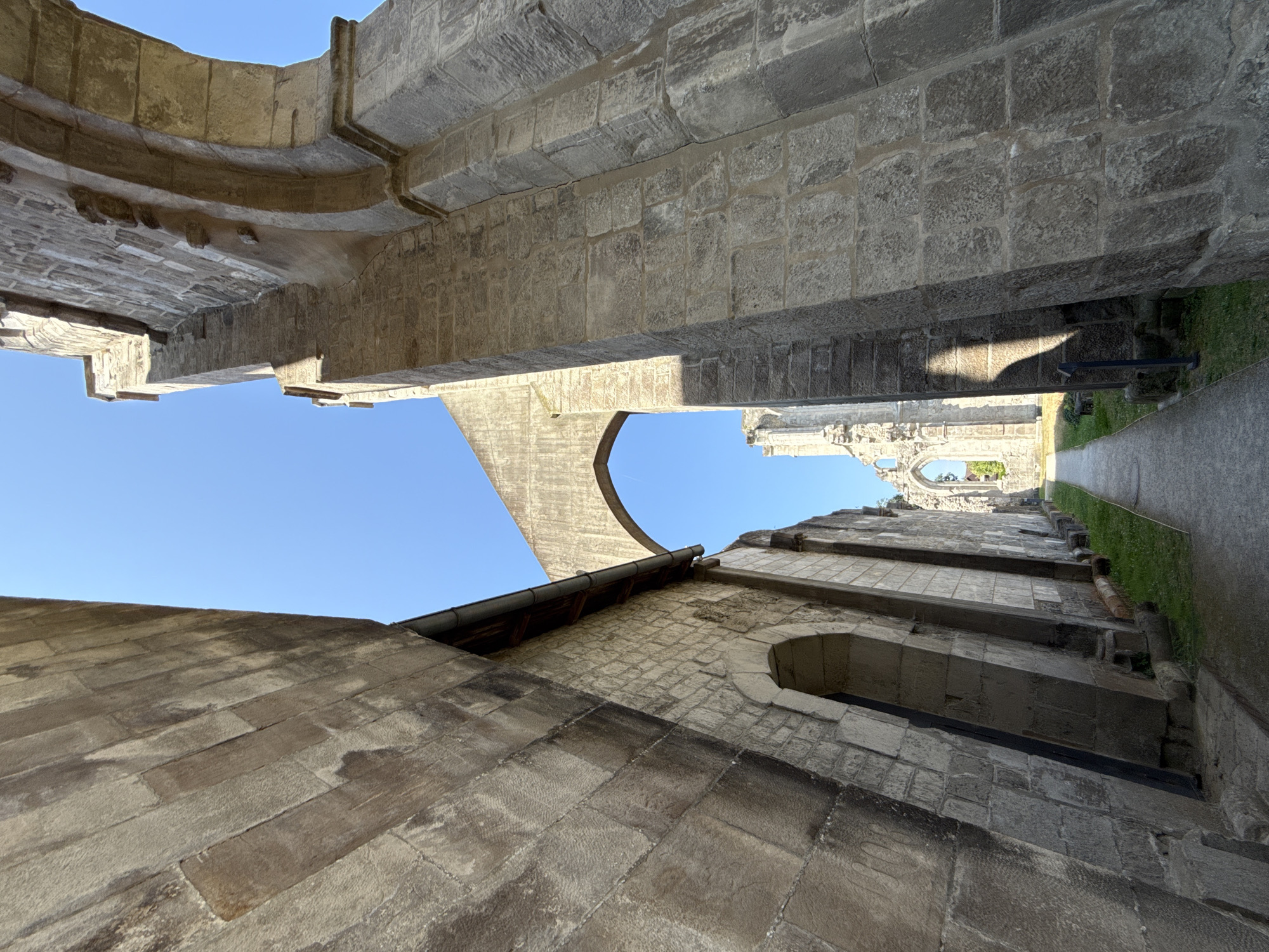 The image shows the exterior of the ruins of the Walkenried Abbey in Walkenried, Germany. The viewpoint is from a pathway looking down a narrow passage between two large stone structures. The stone structures appear to be part of the abbey's architecture, with arched openings and large, rough-hewn stone blocks. The sky above is clear and blue. In the distance, through an archway, more ruins of the abbey are visible, with more arched openings and weathered stone. The pathway is paved with gravel and is bordered by grass. The lighting suggests it is a sunny day, with strong shadows cast by the structures.