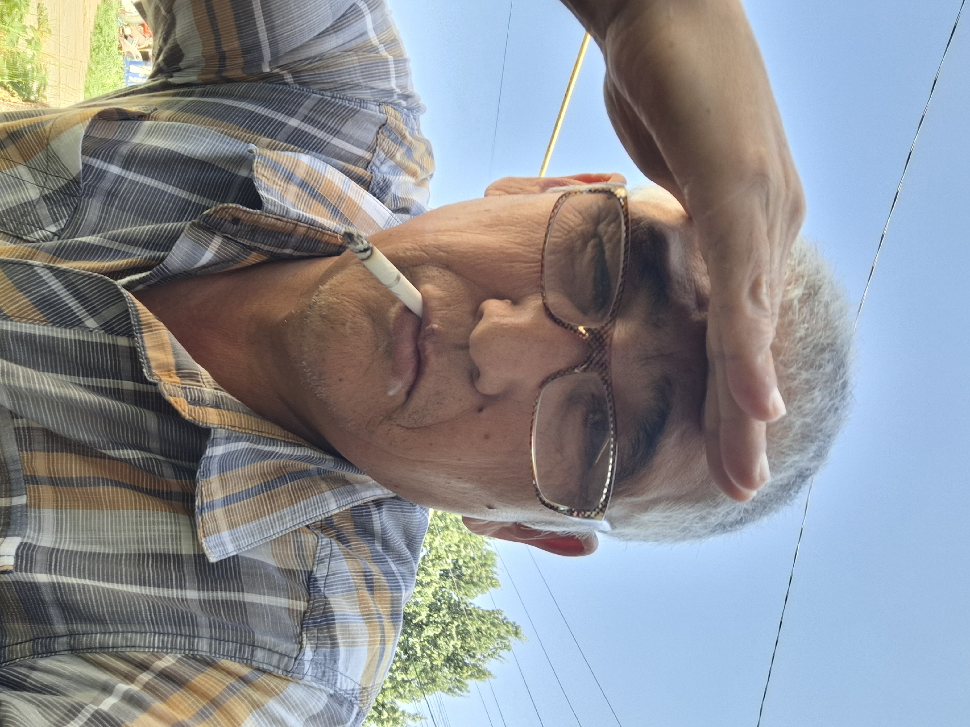 This is a selfie of a middle-aged man with graying hair, wearing glasses and a plaid shirt. He is holding his hand to his forehead as if shielding his eyes from the sun, and smoking a cigarette. The background suggests an outdoor setting with a clear blue sky, power lines, and some trees. The man's facial features are distinct, and the details of his clothing and the cigarette are visible. The image does not provide specific clues that would indicate a location within the city of Almaty, Kazakhstan.