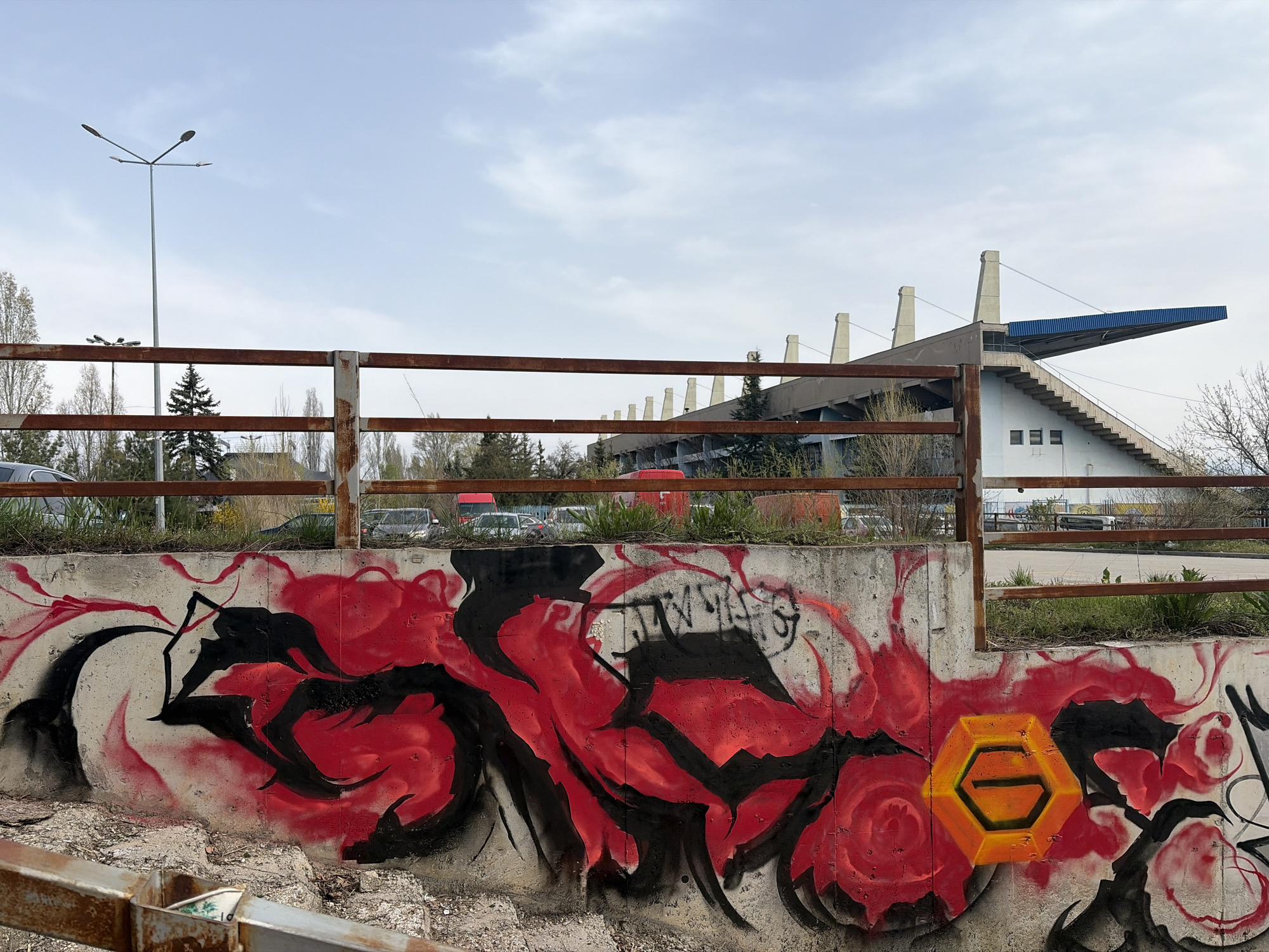 A concrete retaining wall displays vibrant graffiti, predominately in shades of red and black, with a distinct yellow hexagonal figure on the right side. Behind the wall, a rusted metal fence runs horizontally across the mid-ground of the image. Beyond the fence, trees and parked cars are visible. The backdrop features a portion of a stadium with tiered seating, marked by staircases and supported by concrete columns and a blue overhang. A tall street light is present on the left. The sky is overcast. This is a view of the Lokomotiv Stadium in Sofia, Bulgaria.