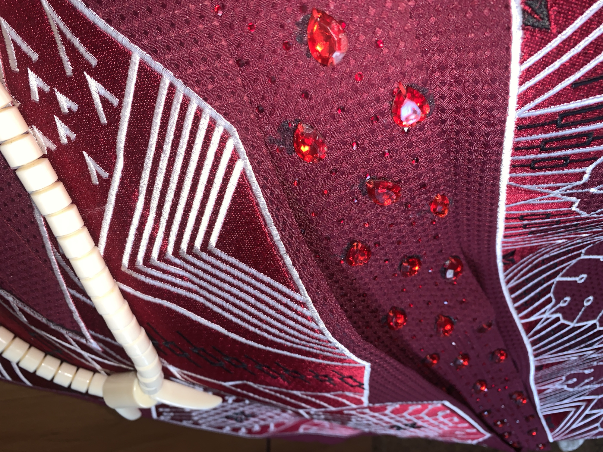 A close-up, shallow-depth-of-field shot captures intricate details of a dark red fabric adorned with various embellishments. Along the top left, a string of cream-colored, cylindrical beads is threaded and draped across the fabric. The fabric itself is textured, appearing to have a subtle raised pattern. A prominent design features thin white lines embroidered in a zigzag or chevron pattern, creating sharp angles and geometric shapes. In another section of the fabric, scattered across the surface, are numerous small, red, faceted gemstones, some larger and more prominent than others. A few larger, teardrop-shaped red gems are also visible, catching the light. The overall impression is one of a richly decorated garment or textile. The image does not contain any information or context related to the city of Gura Top, Nigeria.