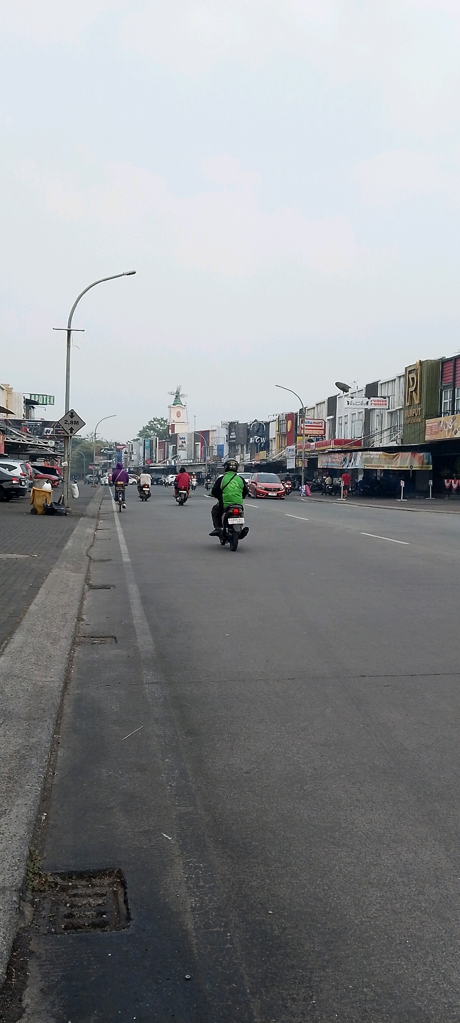 The image depicts a street scene in Cileungsi, Indonesia, likely on a commercial street. In the foreground, a man is riding a motorcycle. He is wearing a green jacket and a black helmet. Several other motorcycles and a bicycle are also on the road. On the left side of the street, there are parked cars, trash cans, and what appears to be a sidewalk with shops or stalls. On the right side of the street, a row of buildings with various signs, including "RUMPUT," "P2W," and "ALFAMART," suggests a commercial area. In the distance, a tall building with a windmill-like structure on top is visible. The sky is overcast.